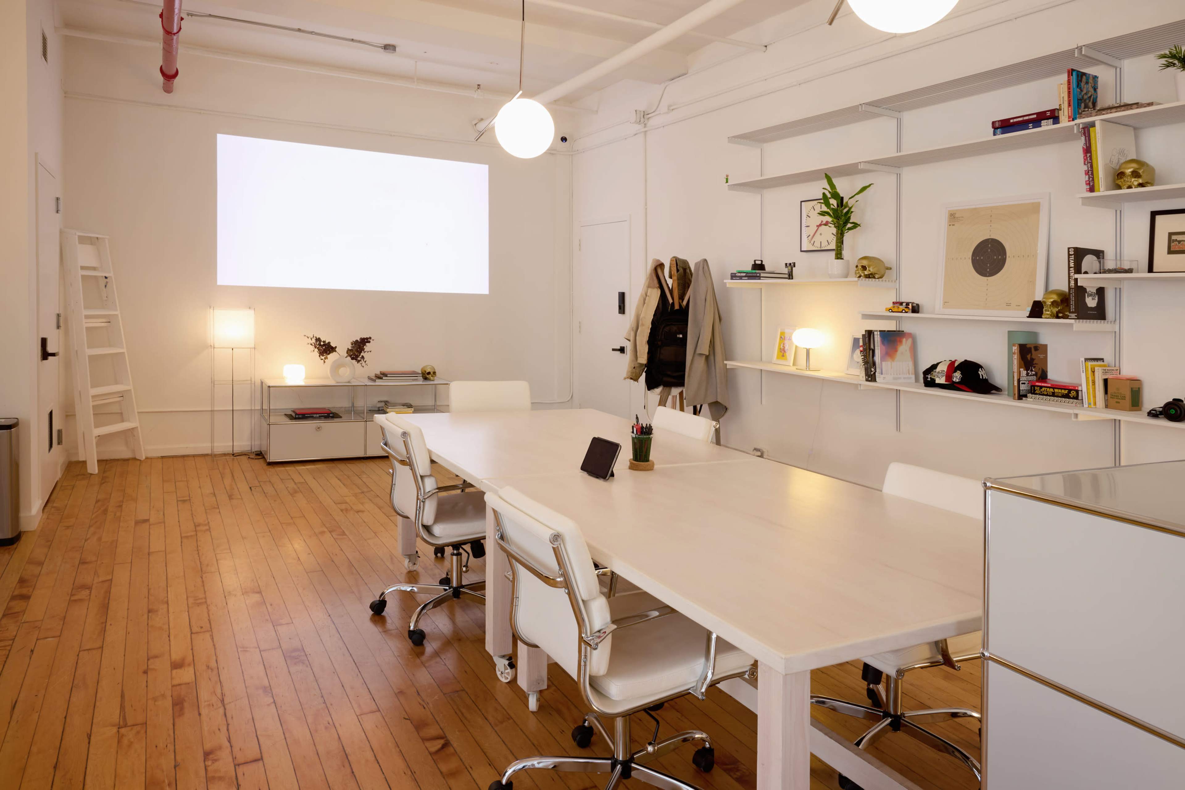 A minimalist conference room features a long white table surrounded by modern chairs, with shelves displaying decor and a projector screen on the wall.