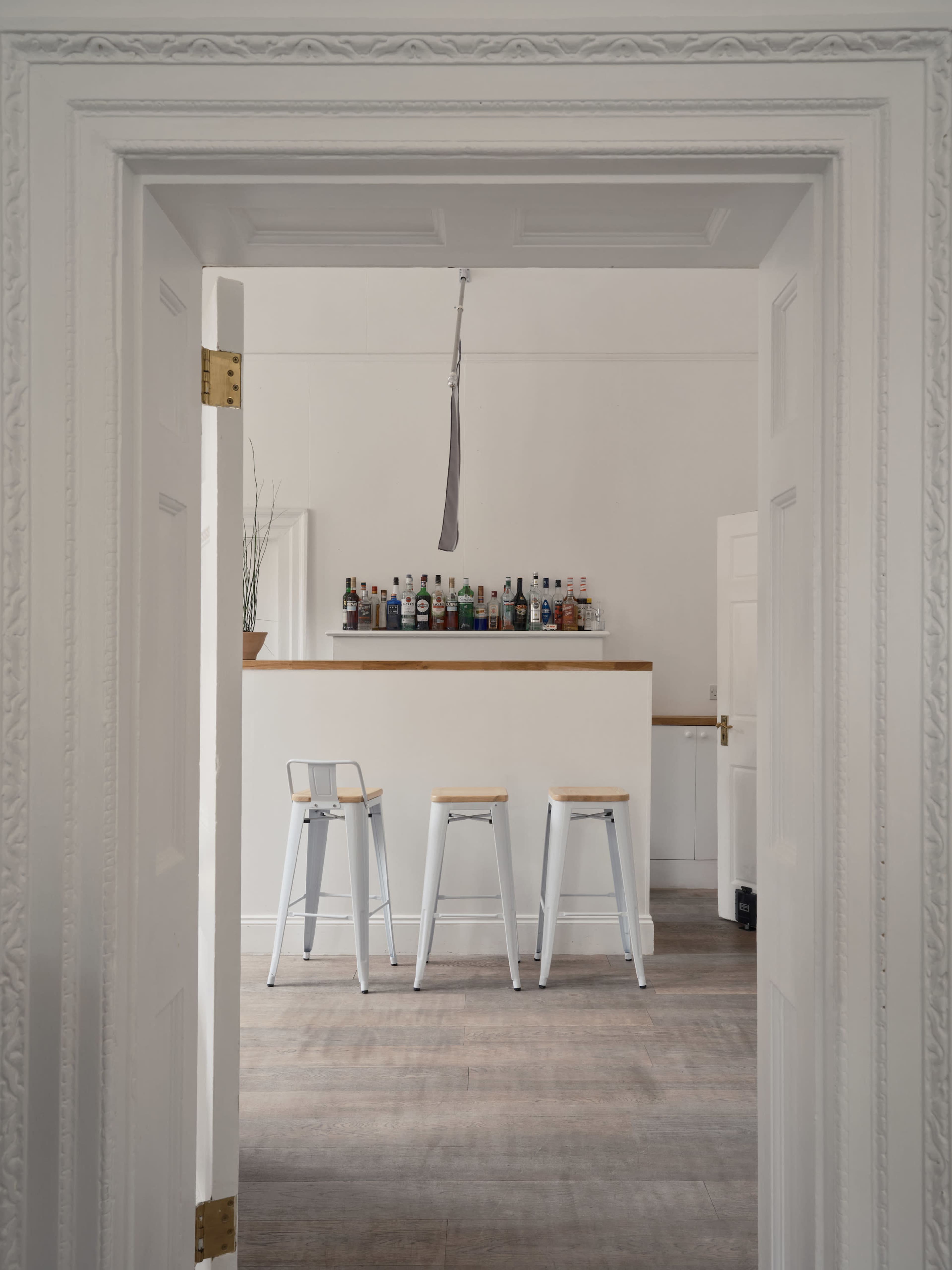 A view through an open door reveals a minimalistic bar area with three high stools and a shelf of bottles in the background.
