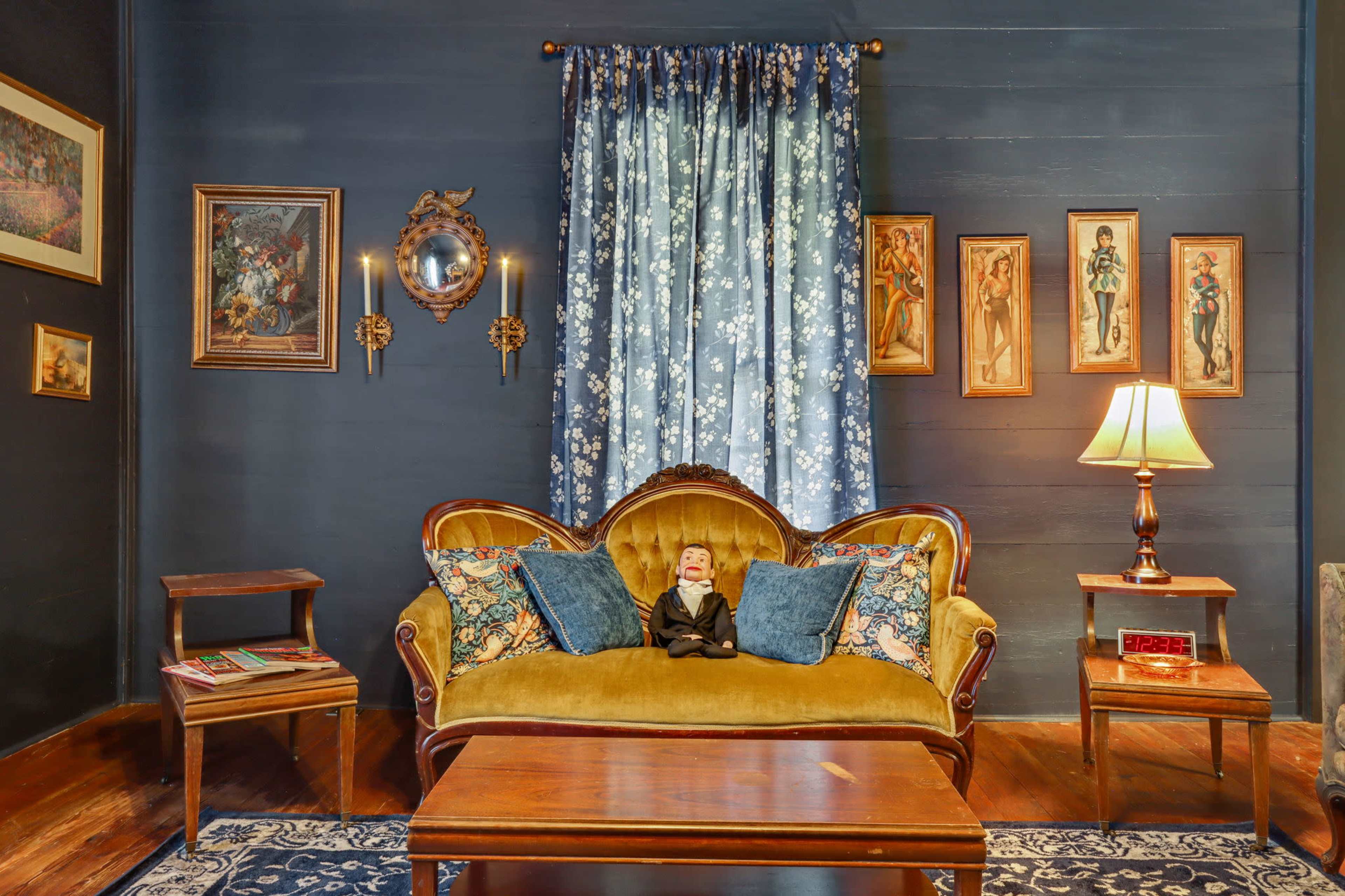 The image shows a vintage living room featuring a yellow velvet sofa adorned with patterned pillows, a wooden coffee table, and walls decorated with framed artwork and a blue floral curtain.