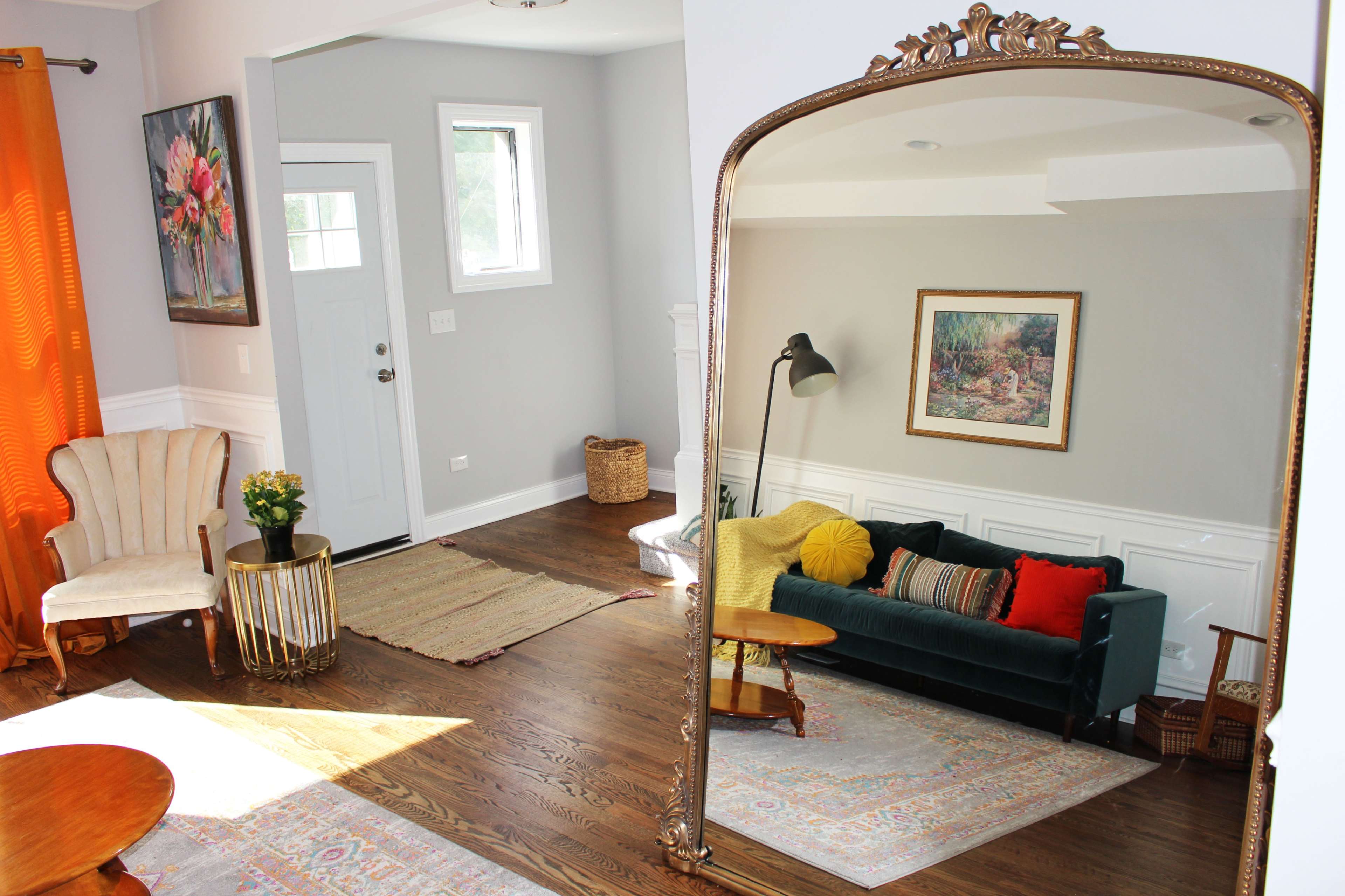 The image shows a living room with a green sofa, two armchairs, a round wooden table, and a large mirror reflecting the space, featuring warm colors and wooden flooring.