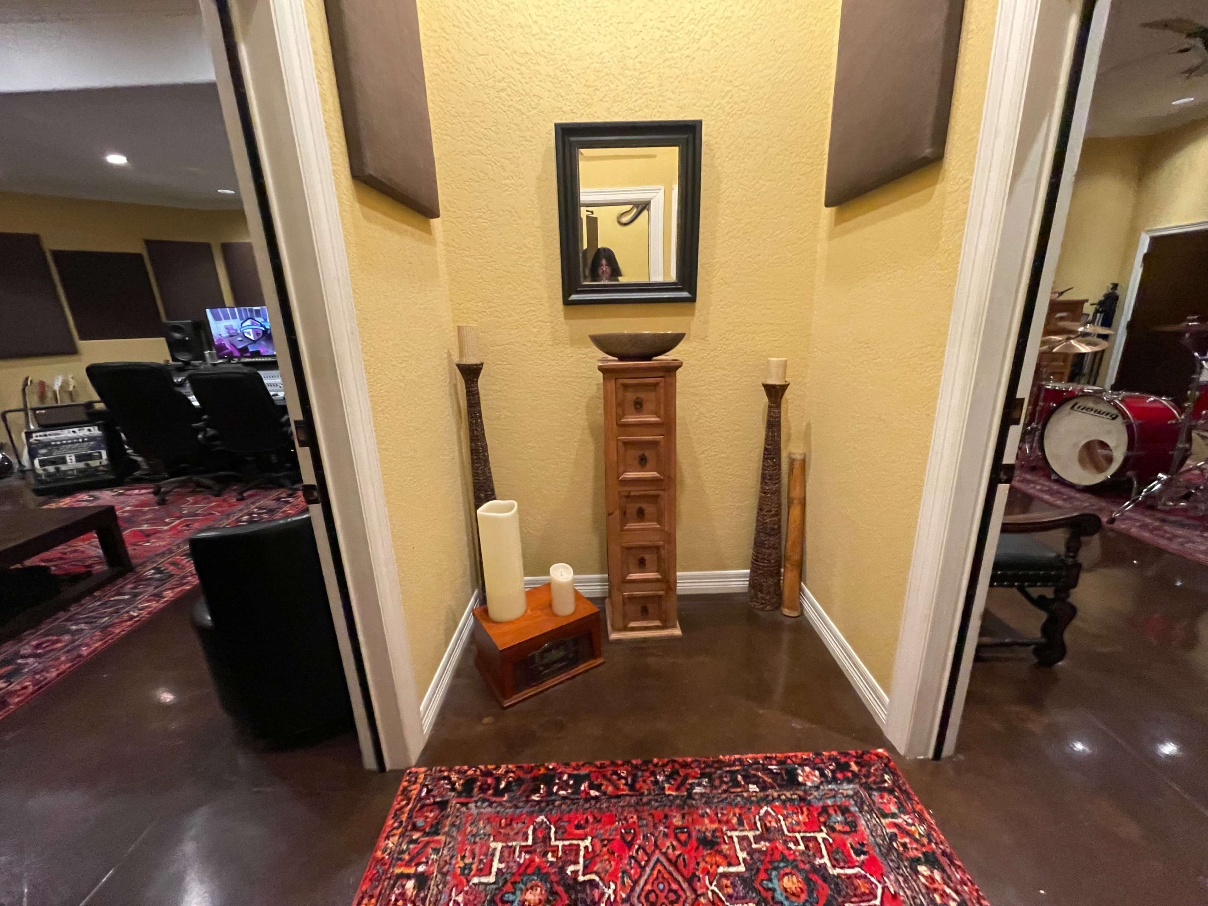 A hallway with yellow walls, decorative items including a mirror, a wooden cabinet, vases, and a red rug, leading to a room with musical equipment.
