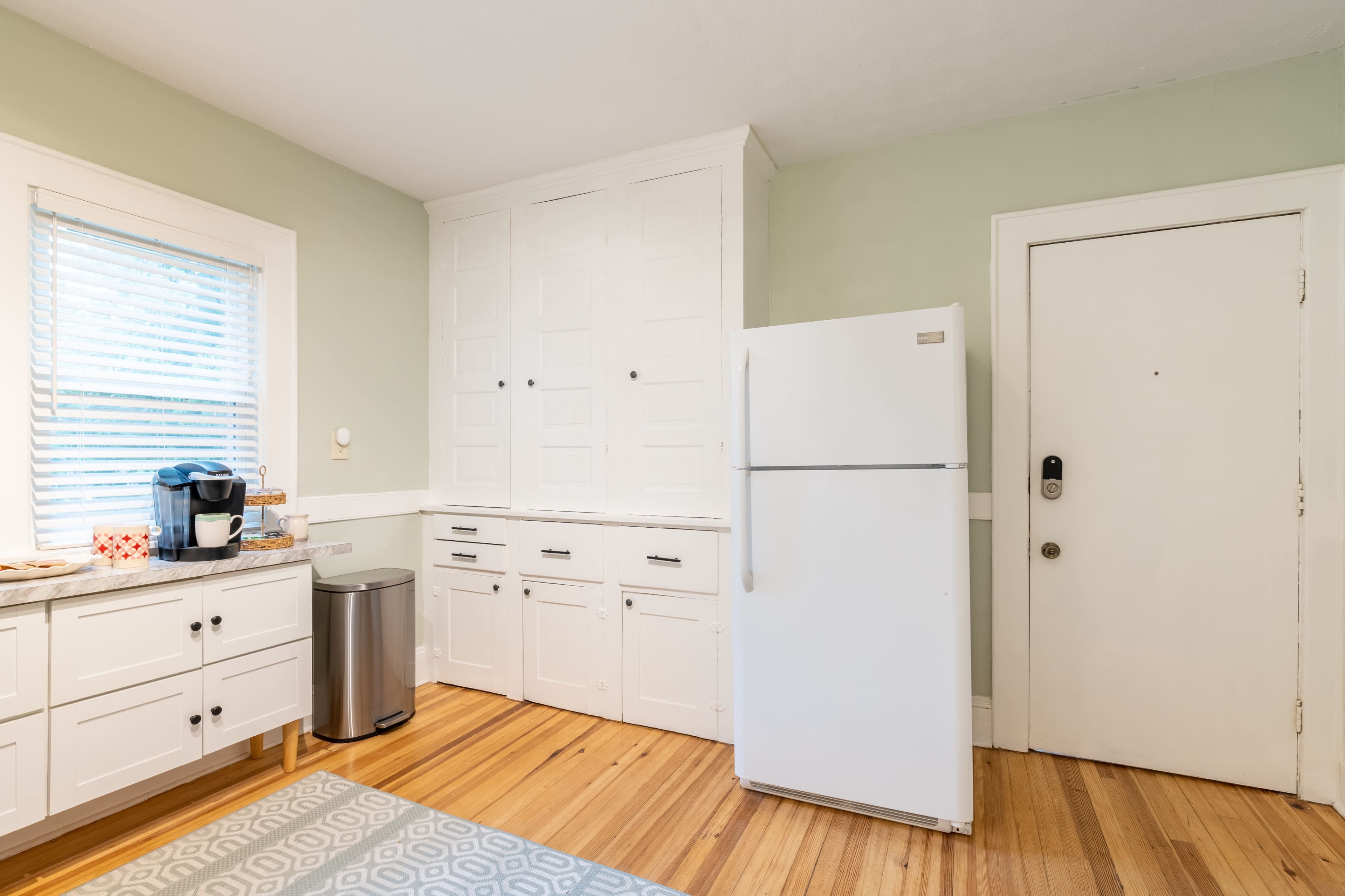 A clean kitchen features white cabinetry, a refrigerator, a coffee maker, and a door leading outside, with hardwood flooring and a patterned rug.