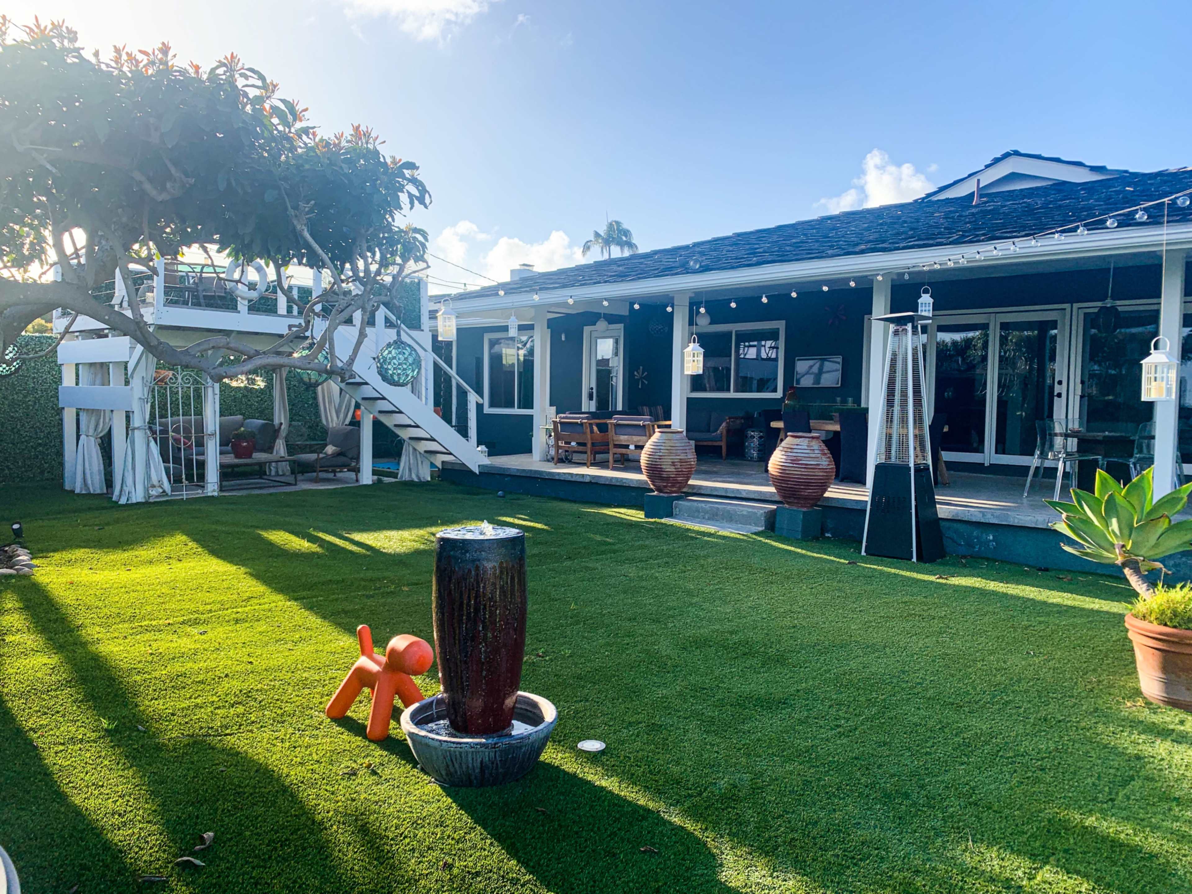 The image shows a well-maintained yard with artificial grass, a patio area with seating, and decorative planters, illuminated by sunlight.
