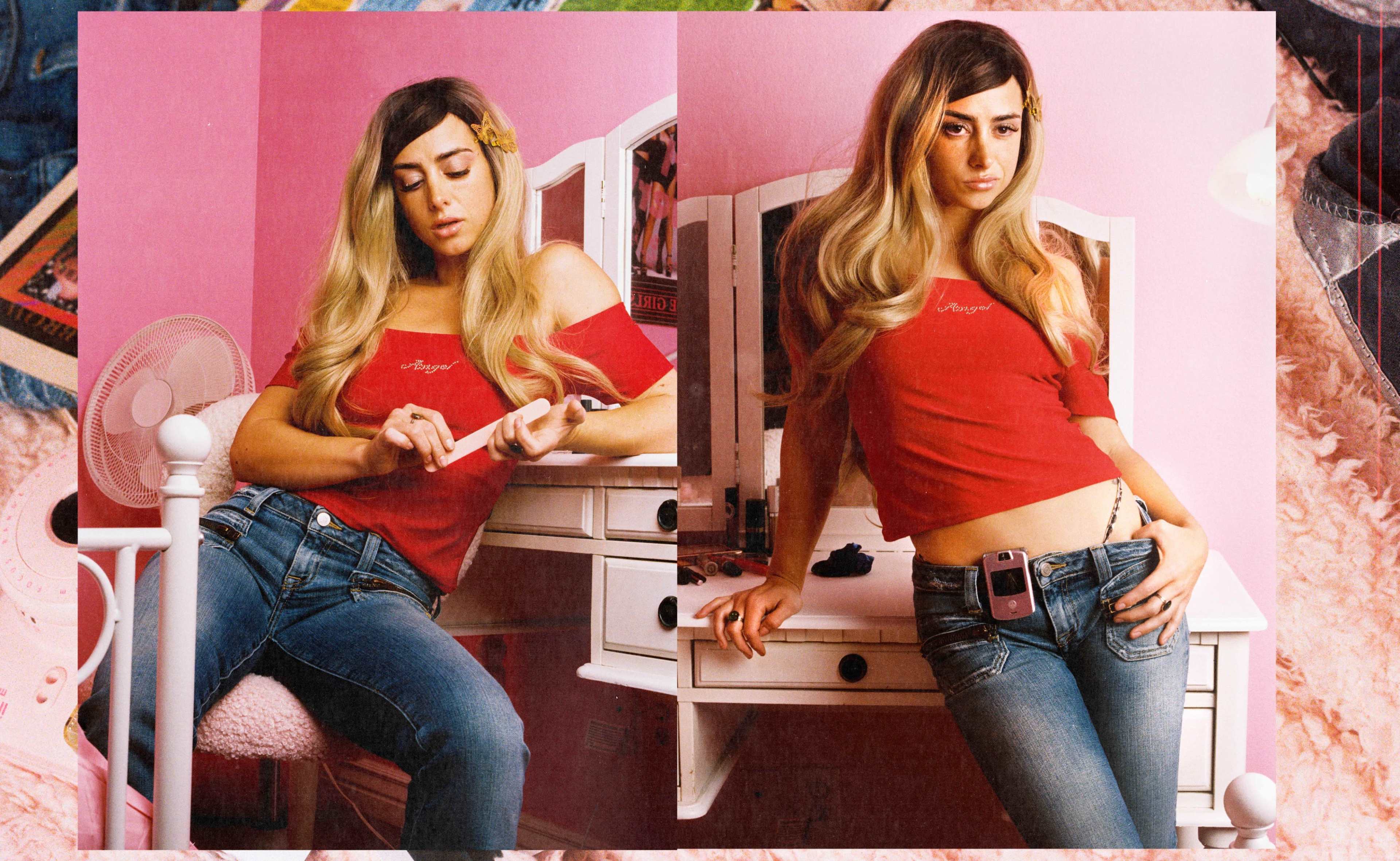 A woman with long hair wearing a red off-the-shoulder top and jeans sits by a vanity in a pink room, posing for the camera.