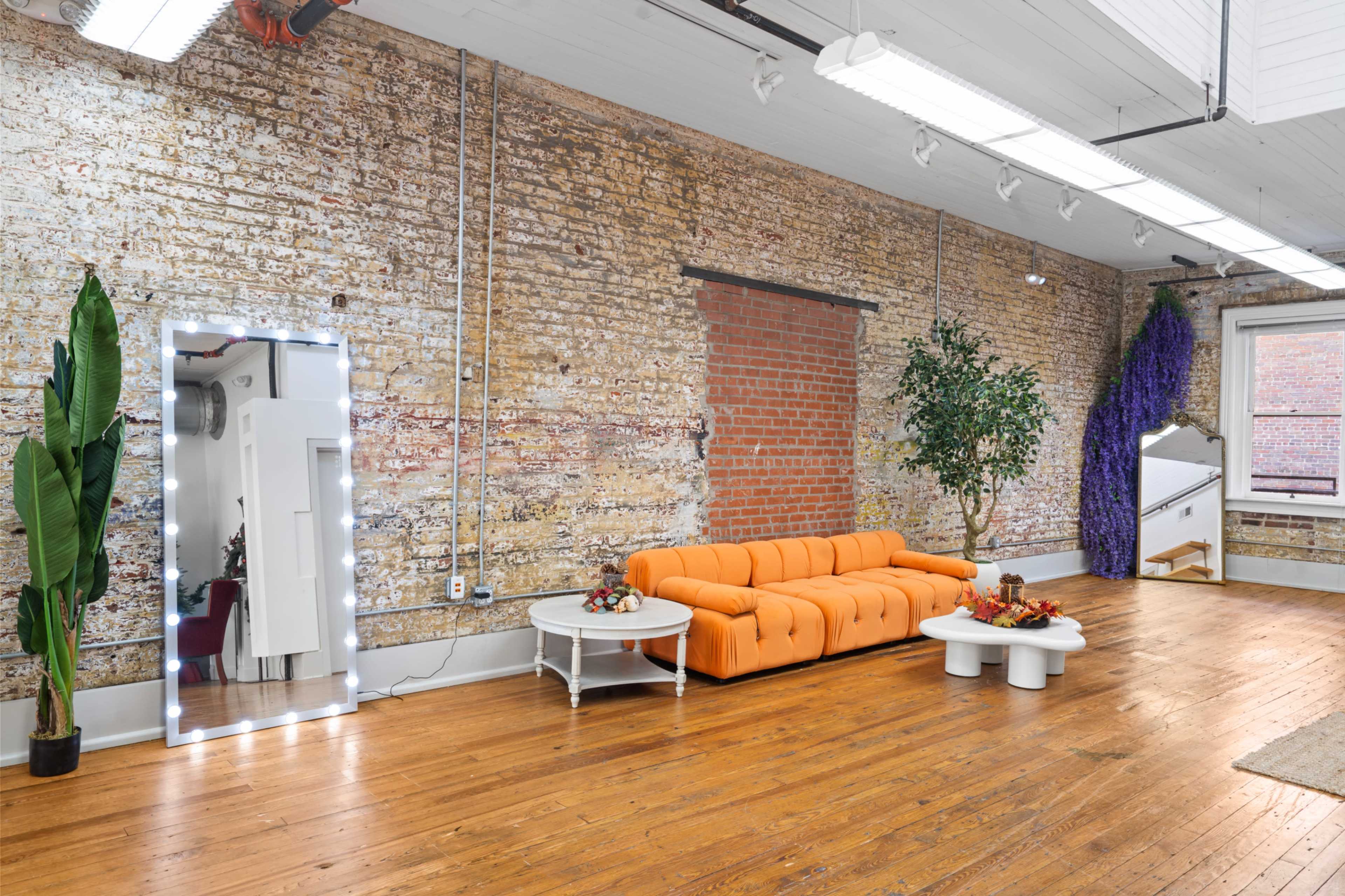 The image shows a spacious room with a textured brick wall, an orange sofa, a mirror with lights, and a small table adorned with decorative items.
