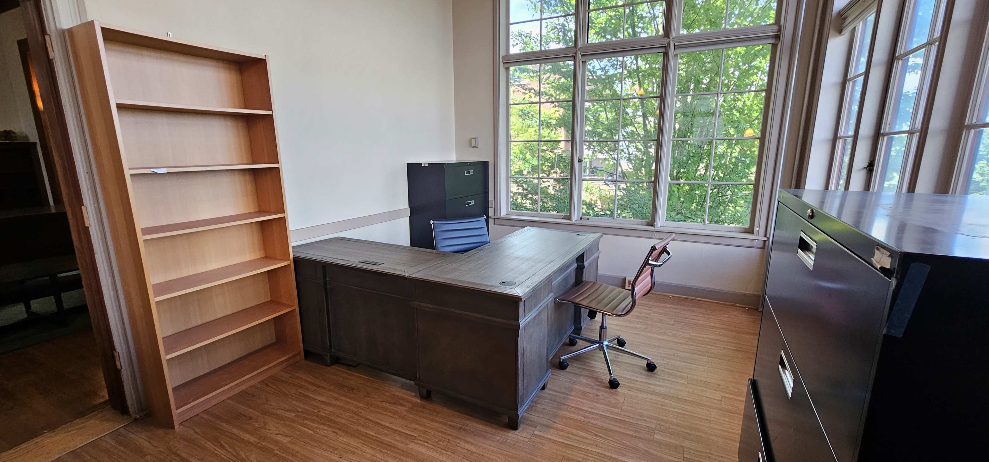 The image shows a vacant office space featuring a desk with a chair, a filing cabinet, and a bookshelf against the wall, with large windows allowing natural light to enter.