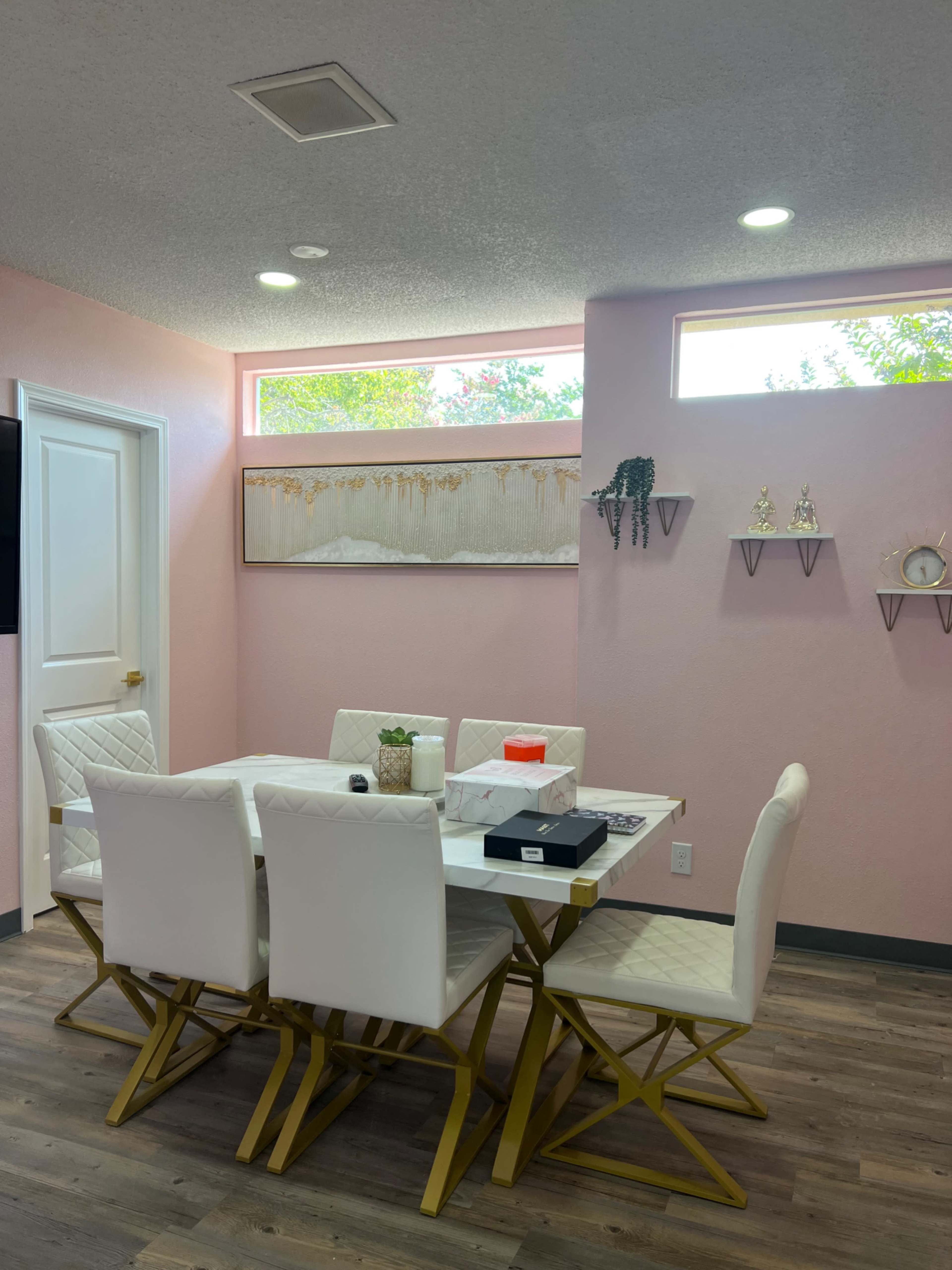 A dining table with white chairs and a gold base is positioned in a brightly lit room with pink walls and windows, featuring decorative wall shelves and artwork.