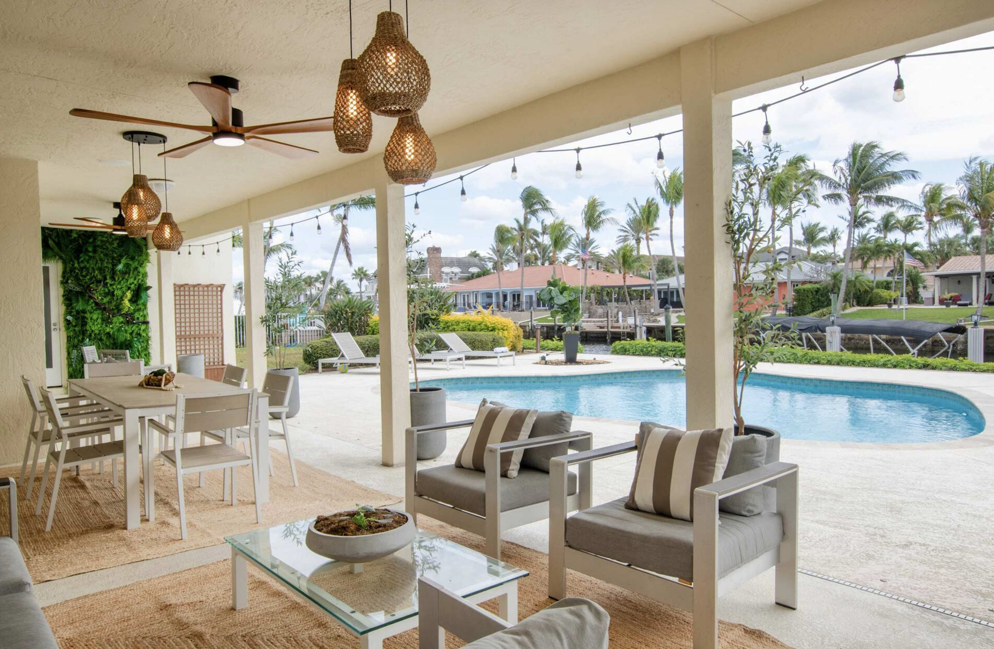 The image shows a covered outdoor patio area with modern furniture, a dining table, and a swimming pool surrounded by palm trees and residential buildings in the background.