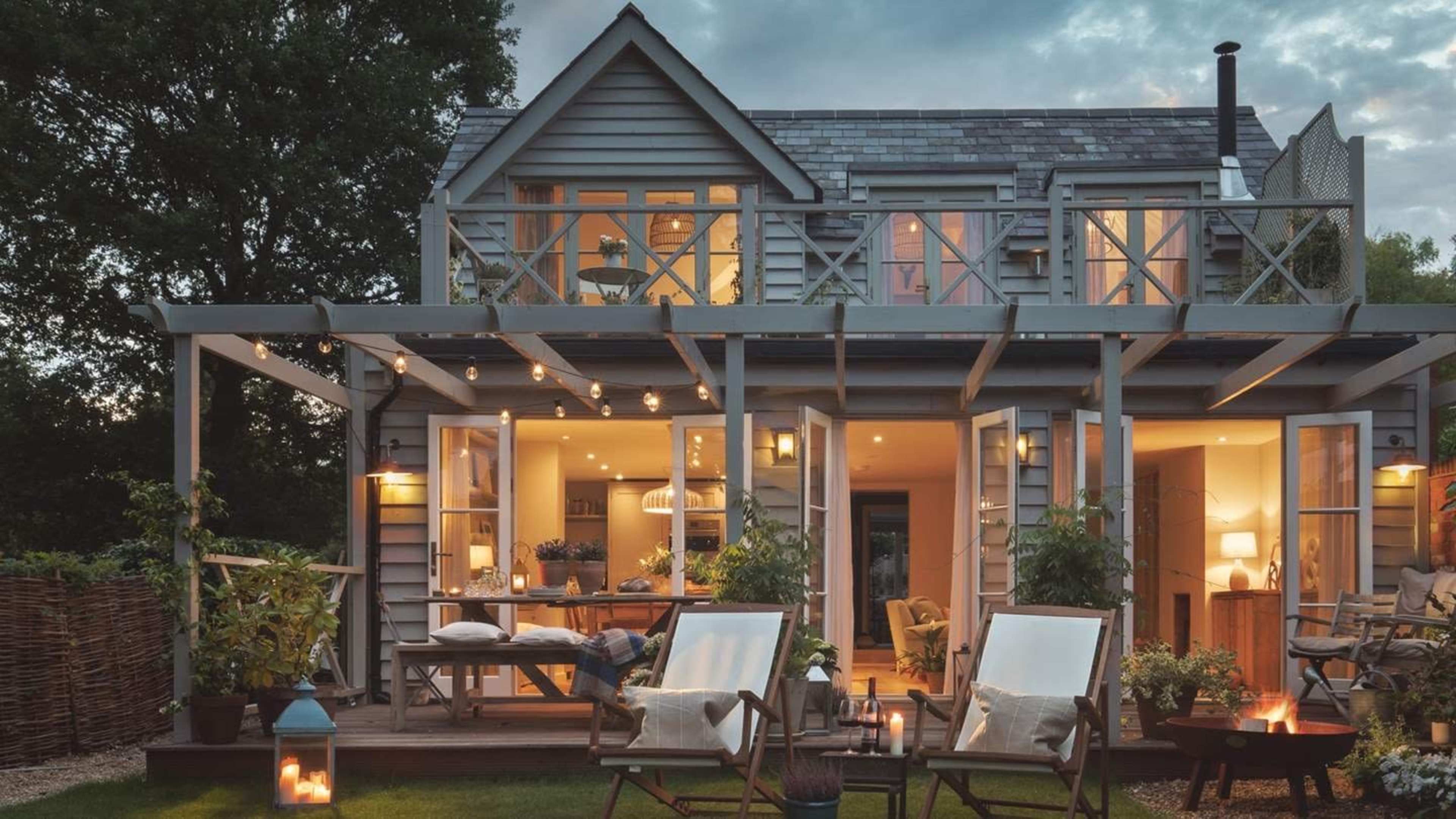A cozy house with a deck and patio area is illuminated by outdoor lights, surrounded by greenery and featuring large windows that reveal a warm interior.
