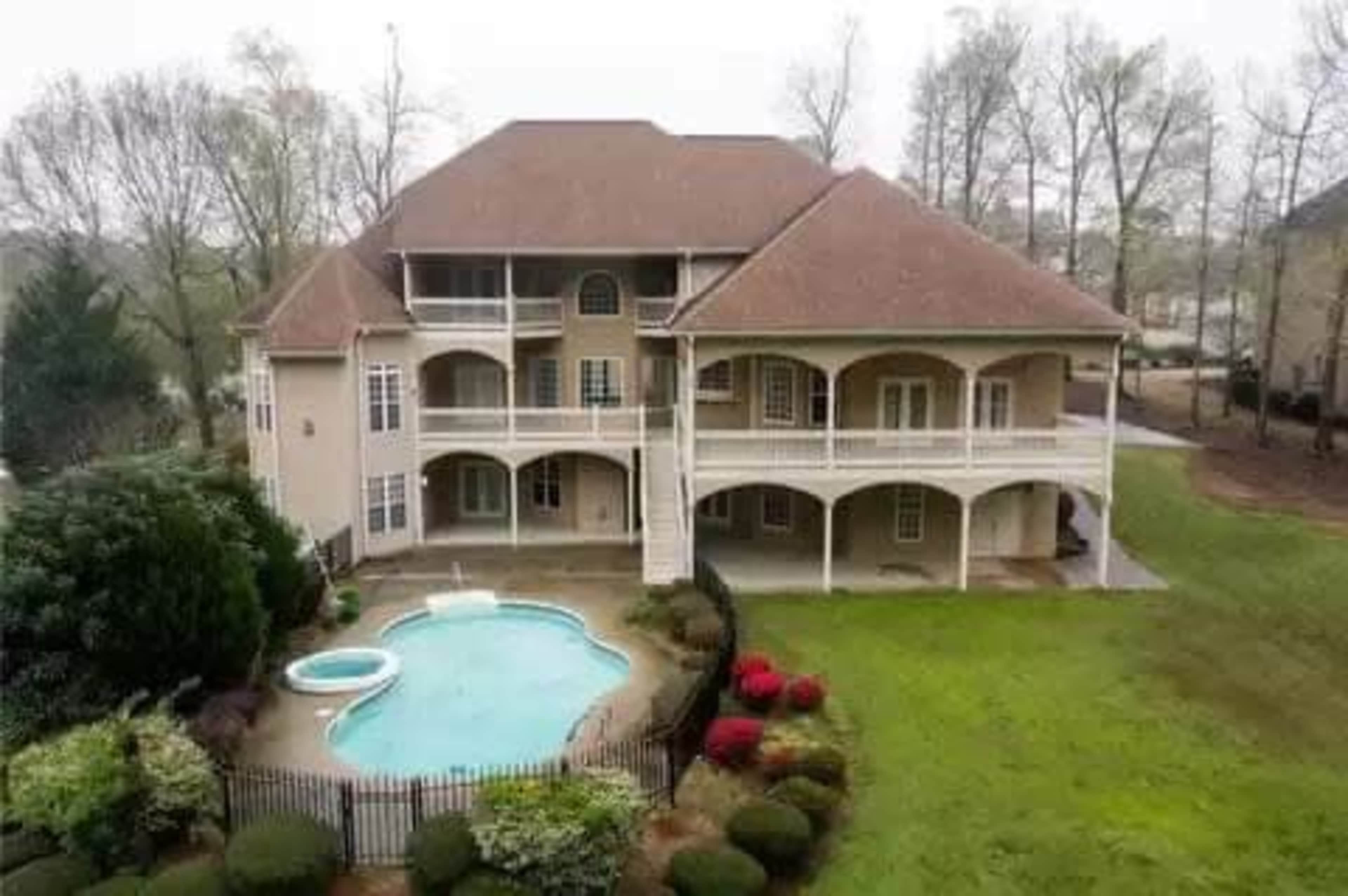 A large two-story house with a brown roof features a fenced-in backyard with a swimming pool and landscaped greenery.