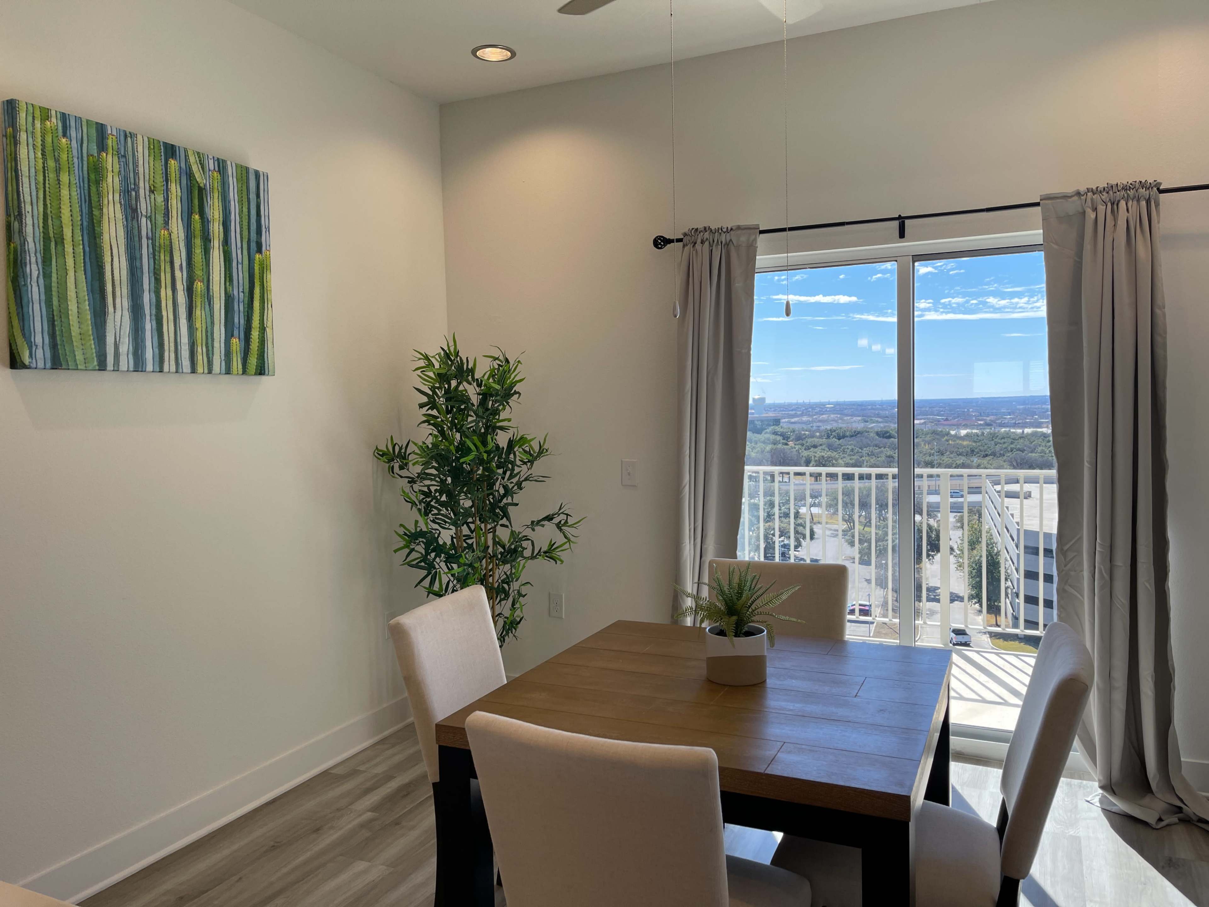 A dining area features a wooden table with four chairs, a potted plant, and a large window offering a view of the outdoors.