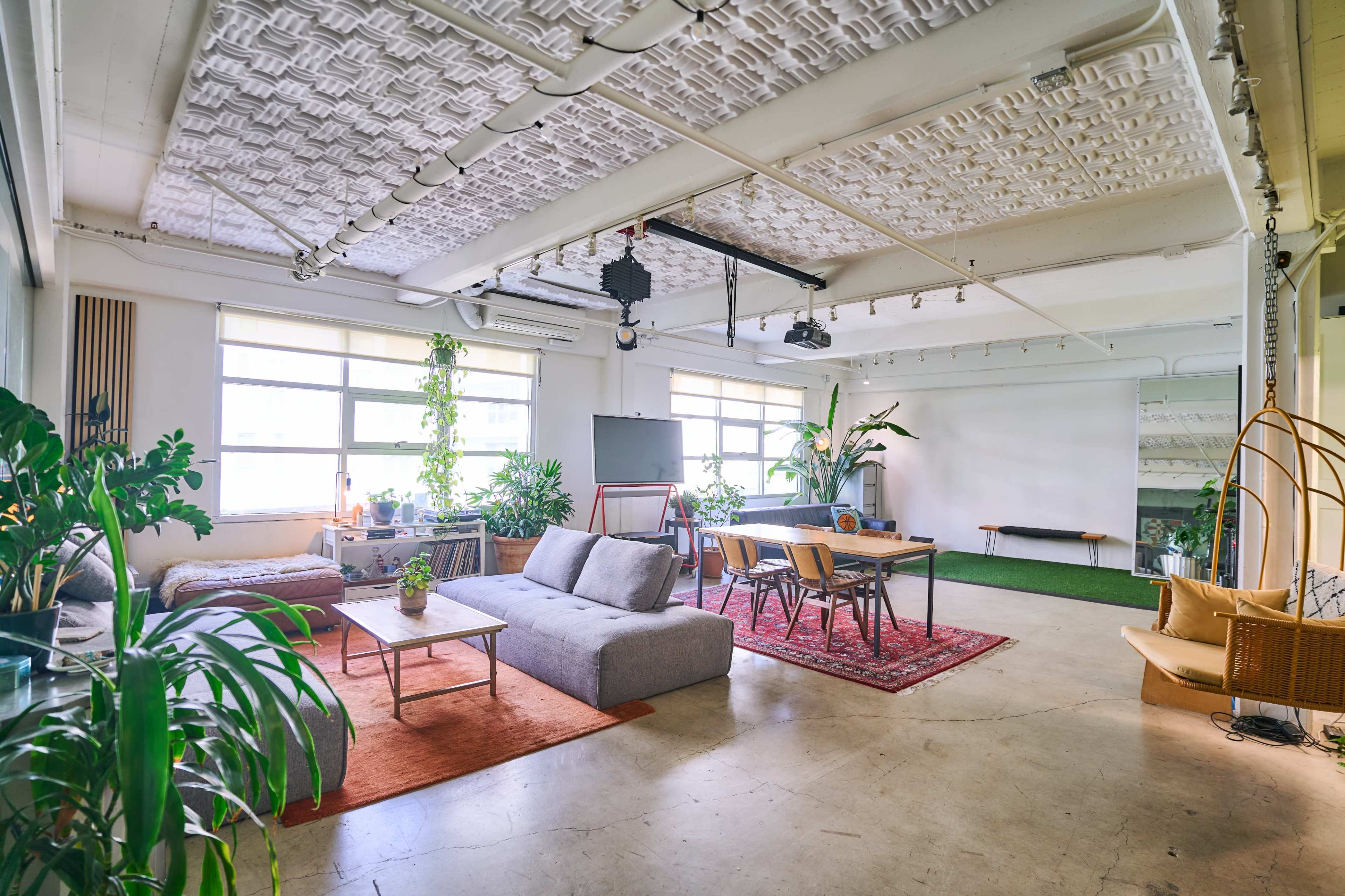 The image shows a spacious, modern workspace with large windows, various plants, a combination of seating areas, and a dining table.
