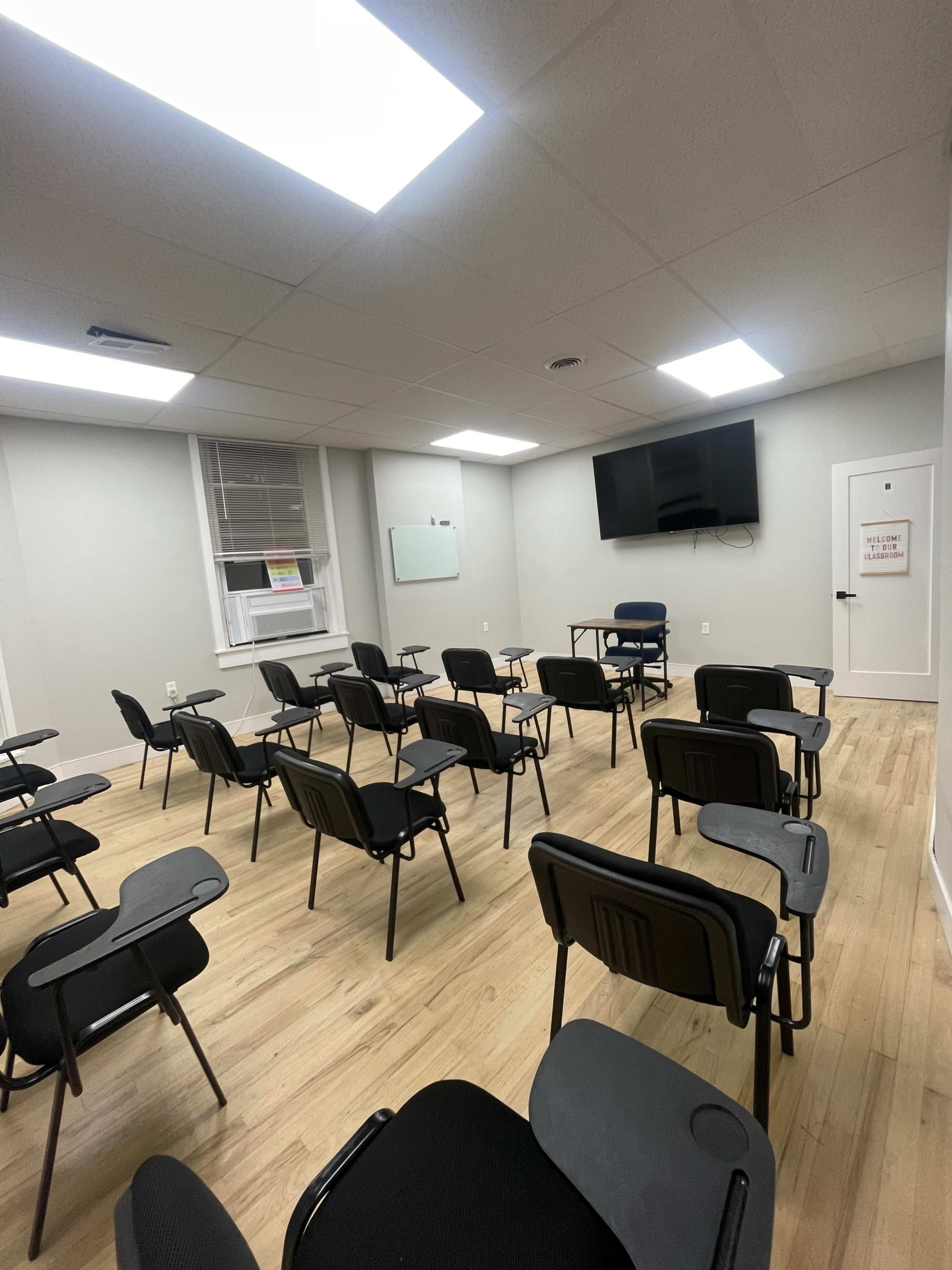 The image shows a sparsely furnished room with rows of black chairs facing a table and a television screen mounted on the wall.