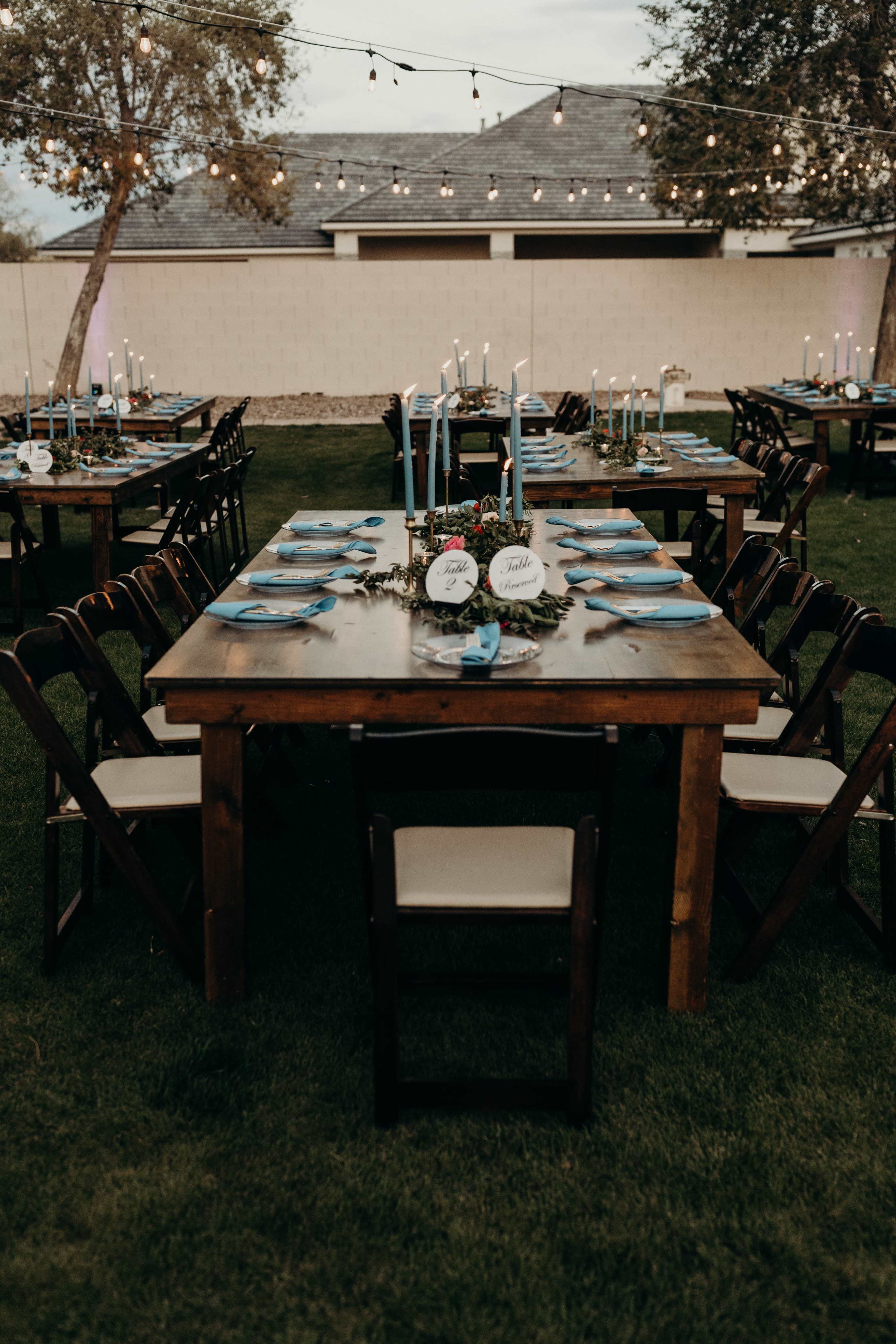 A long wooden table is set for a dinner gathering outdoors, surrounded by chairs, with candles and blue plates arranged atop it, and string lights overhead.