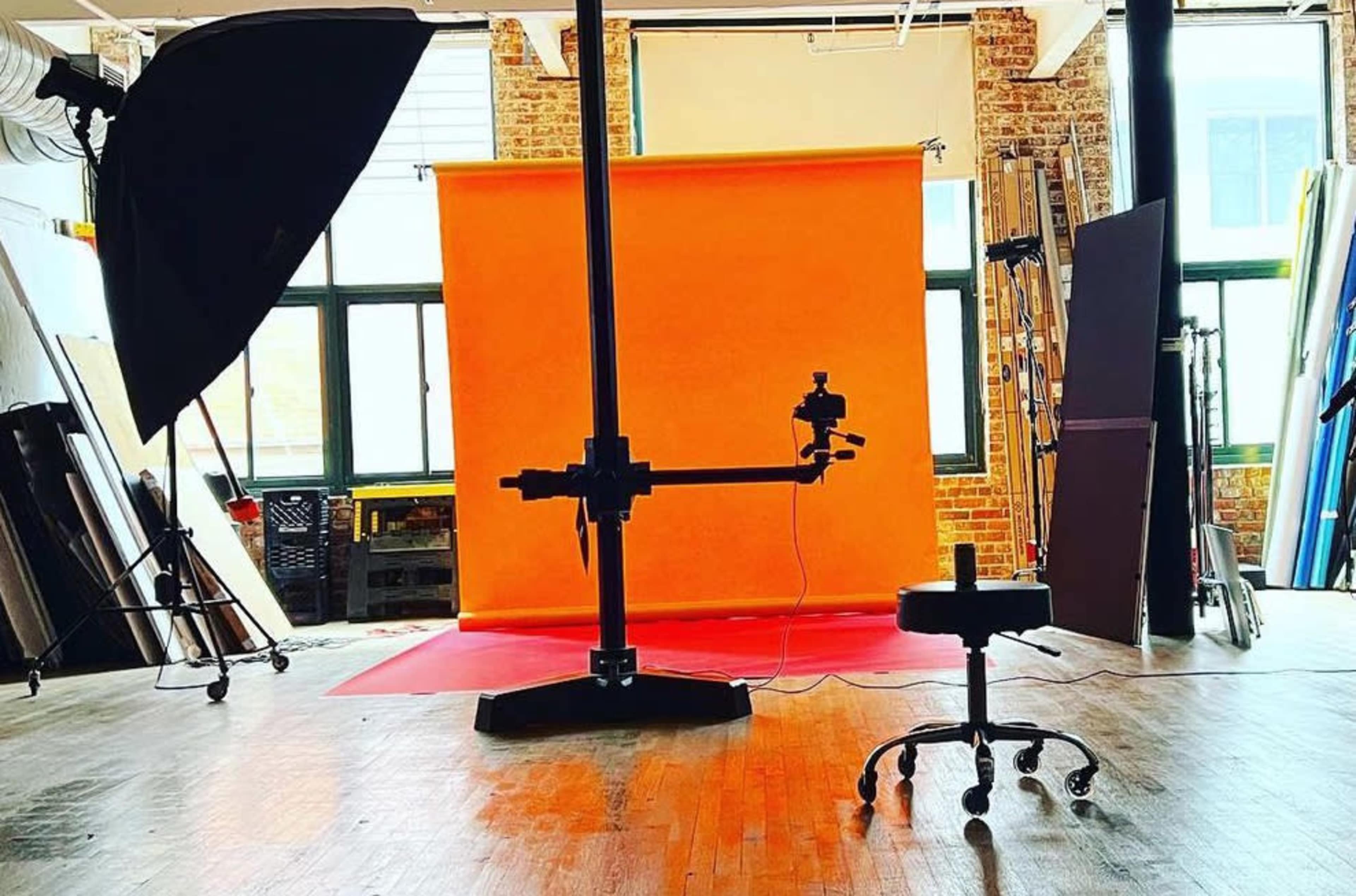 A photography studio features a bright orange backdrop, a lighting setup, and a swivel chair on a wooden floor.