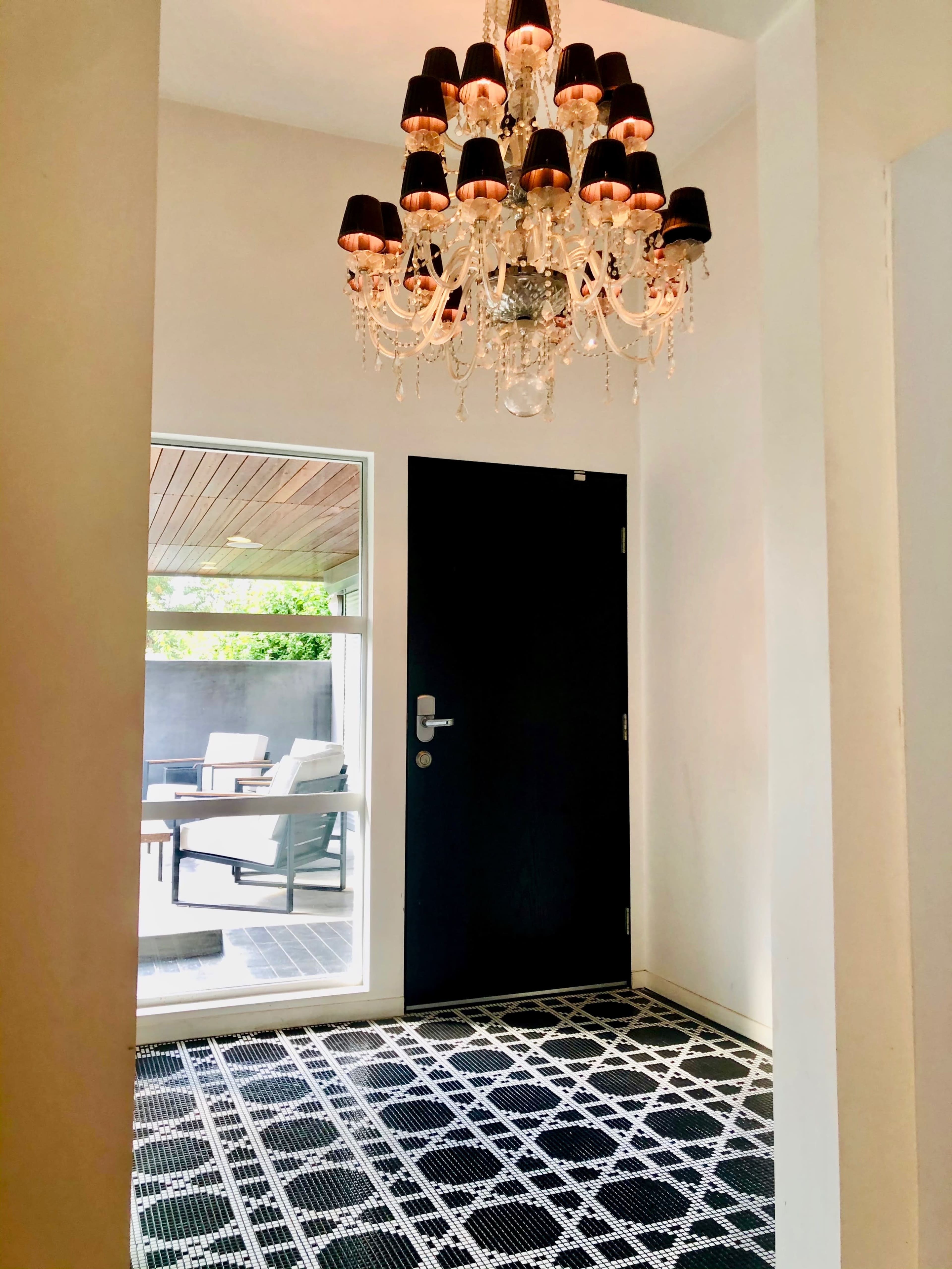 The image shows an entryway featuring a black door, a chandelier with shades, and a patterned black and white floor, leading to an outdoor seating area through a large glass door.