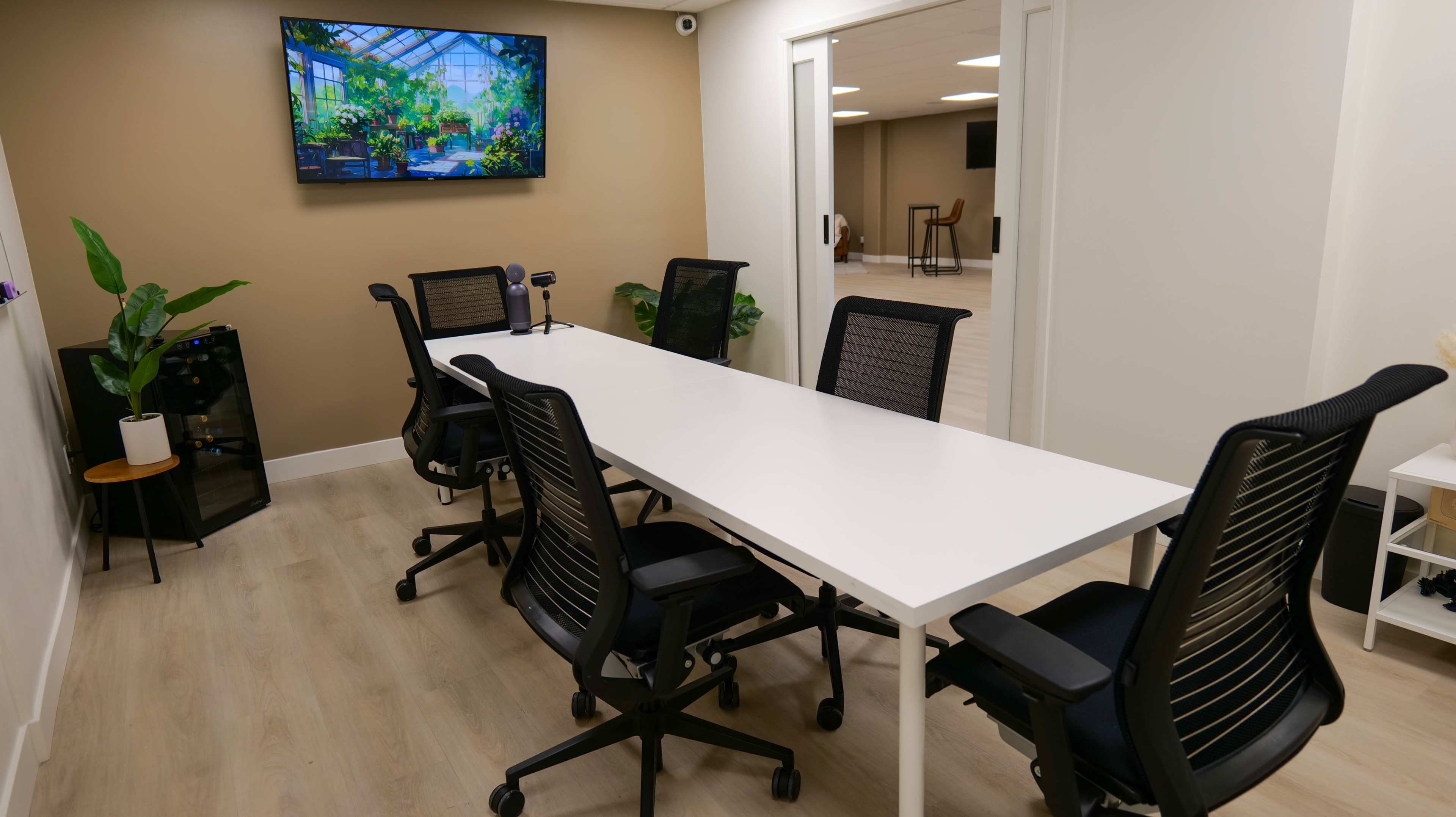 A modern conference room features a long white table surrounded by black ergonomic chairs, with a large screen displaying greenery on the wall.