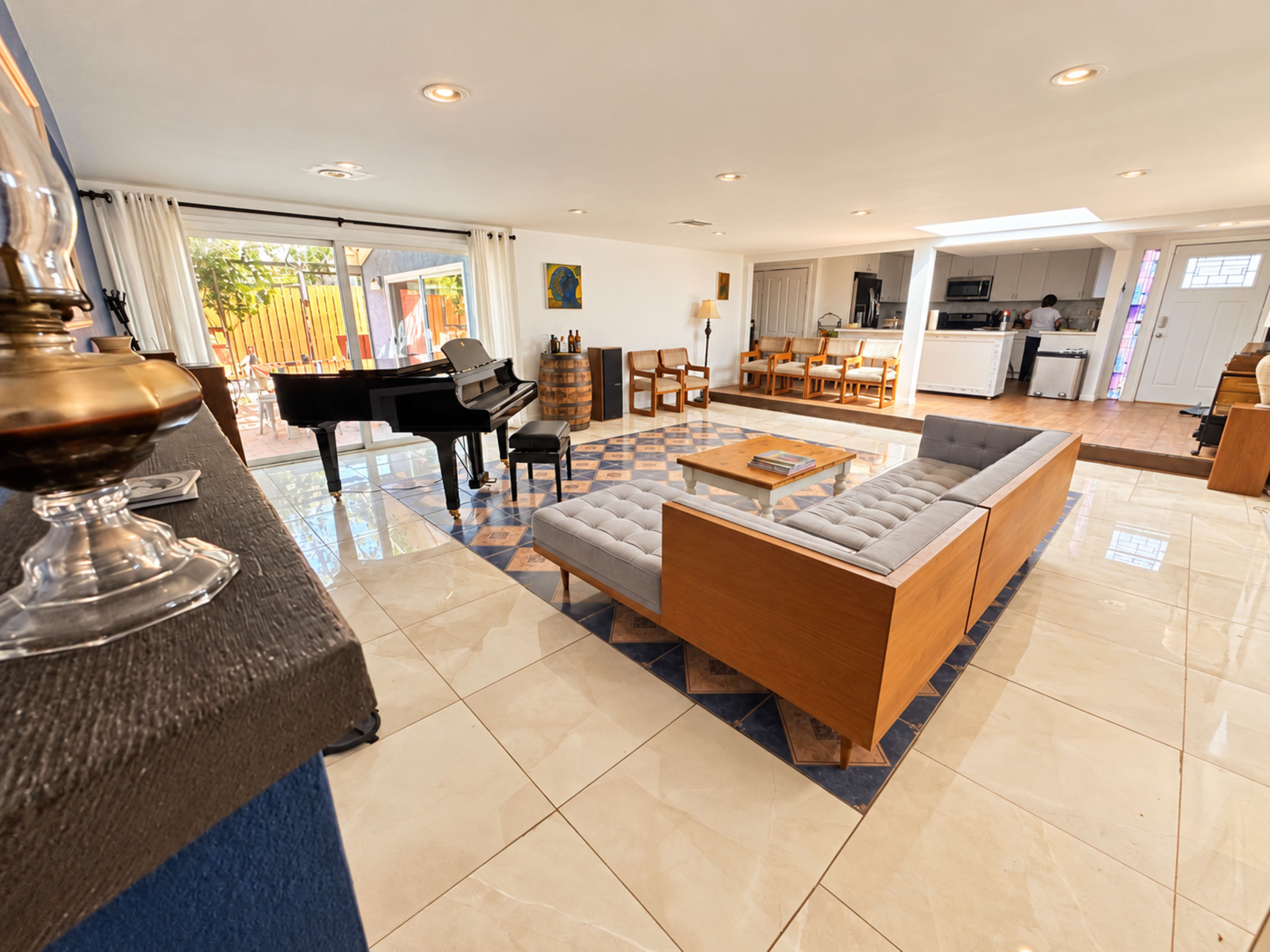 The image shows a spacious living area featuring a grand piano, a modern couch, and a dining setup in an open layout with natural light flowing in through large windows.