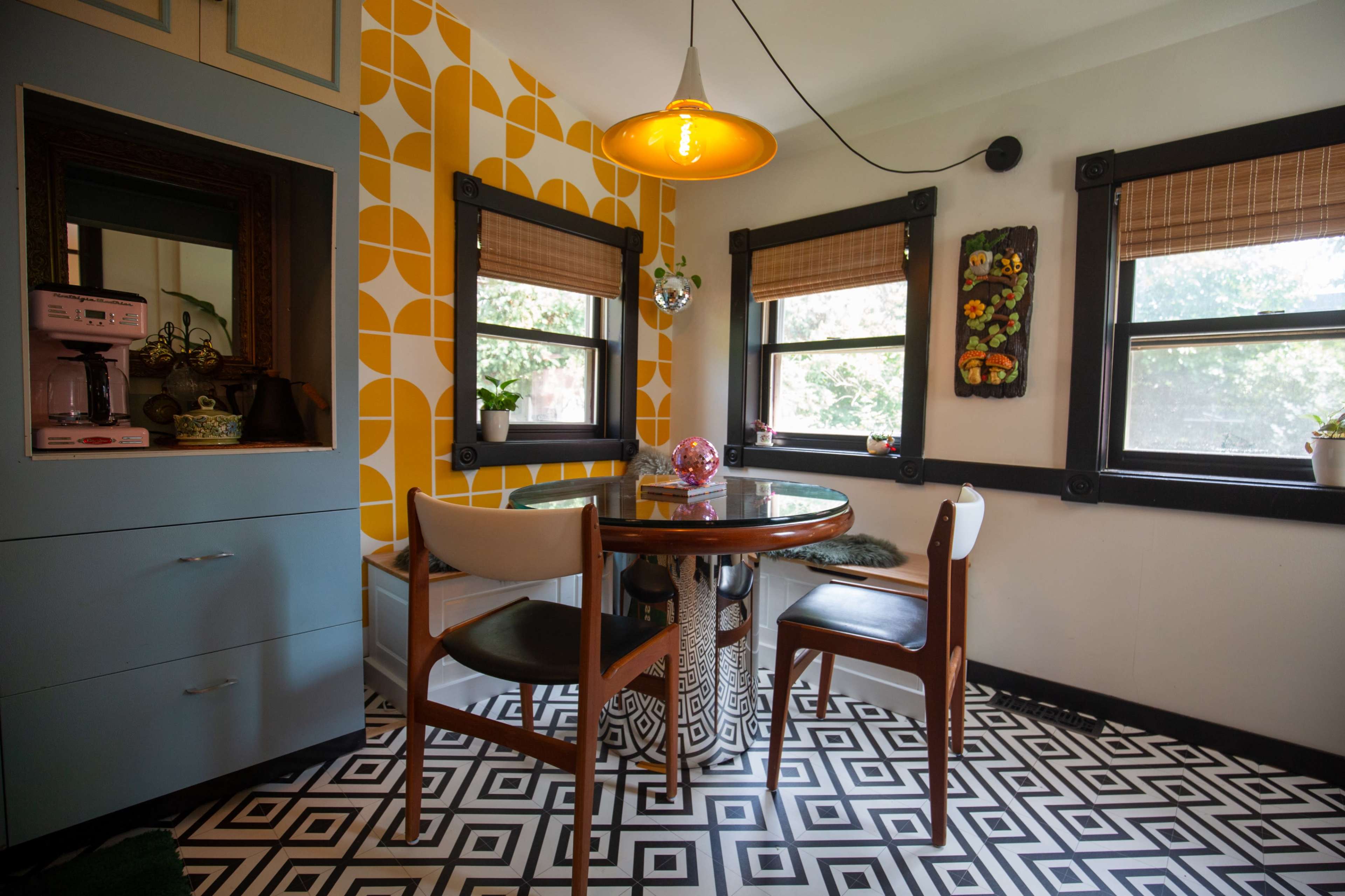A dining area features a round table surrounded by four chairs, patterned flooring, and walls with geometric wallpaper and windows adorned with bamboo shades.
