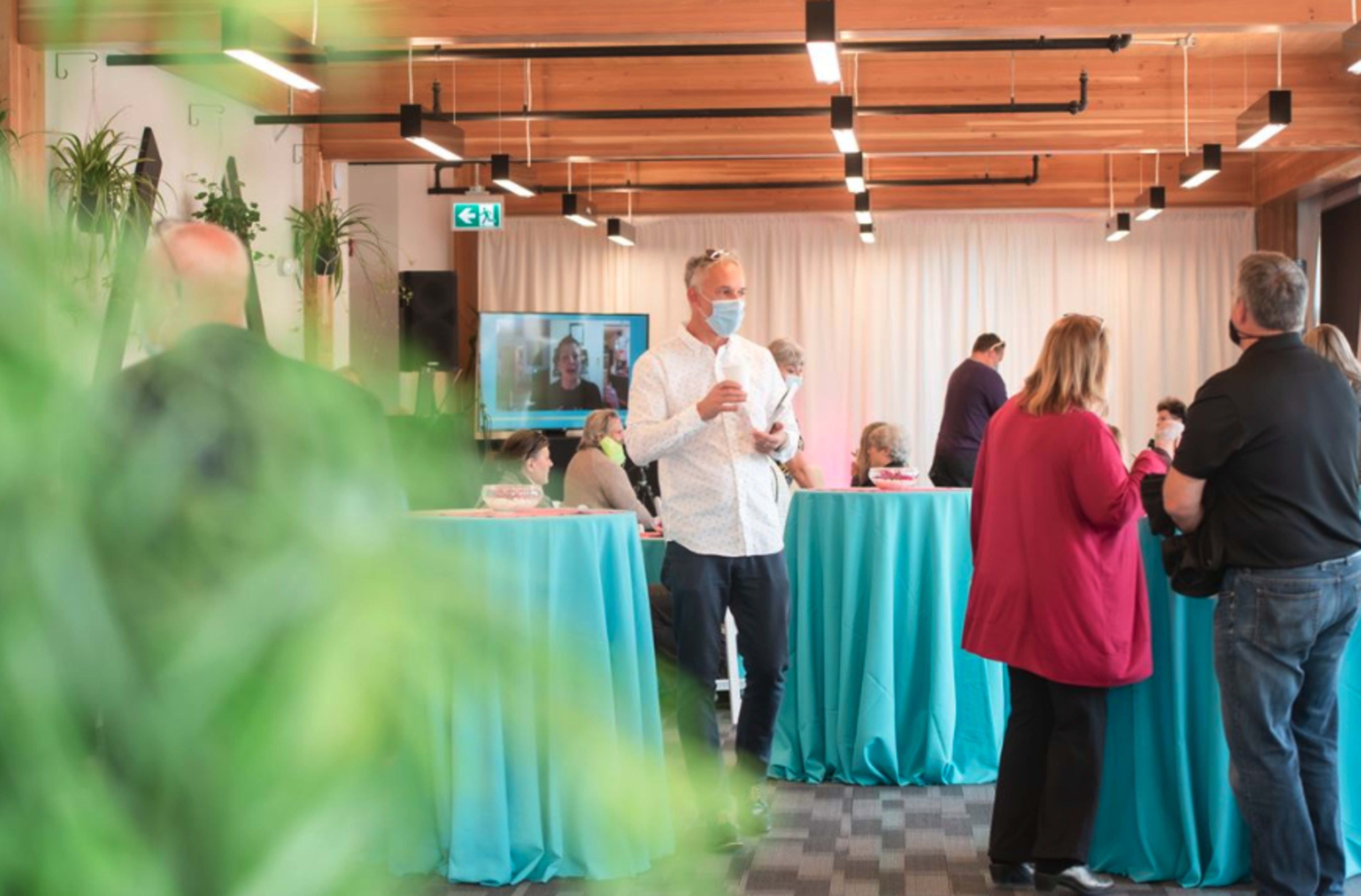 A group of people stands around teal tables in a room with wooden beams, some engaging in conversation while others watch a video presentation on a screen.
