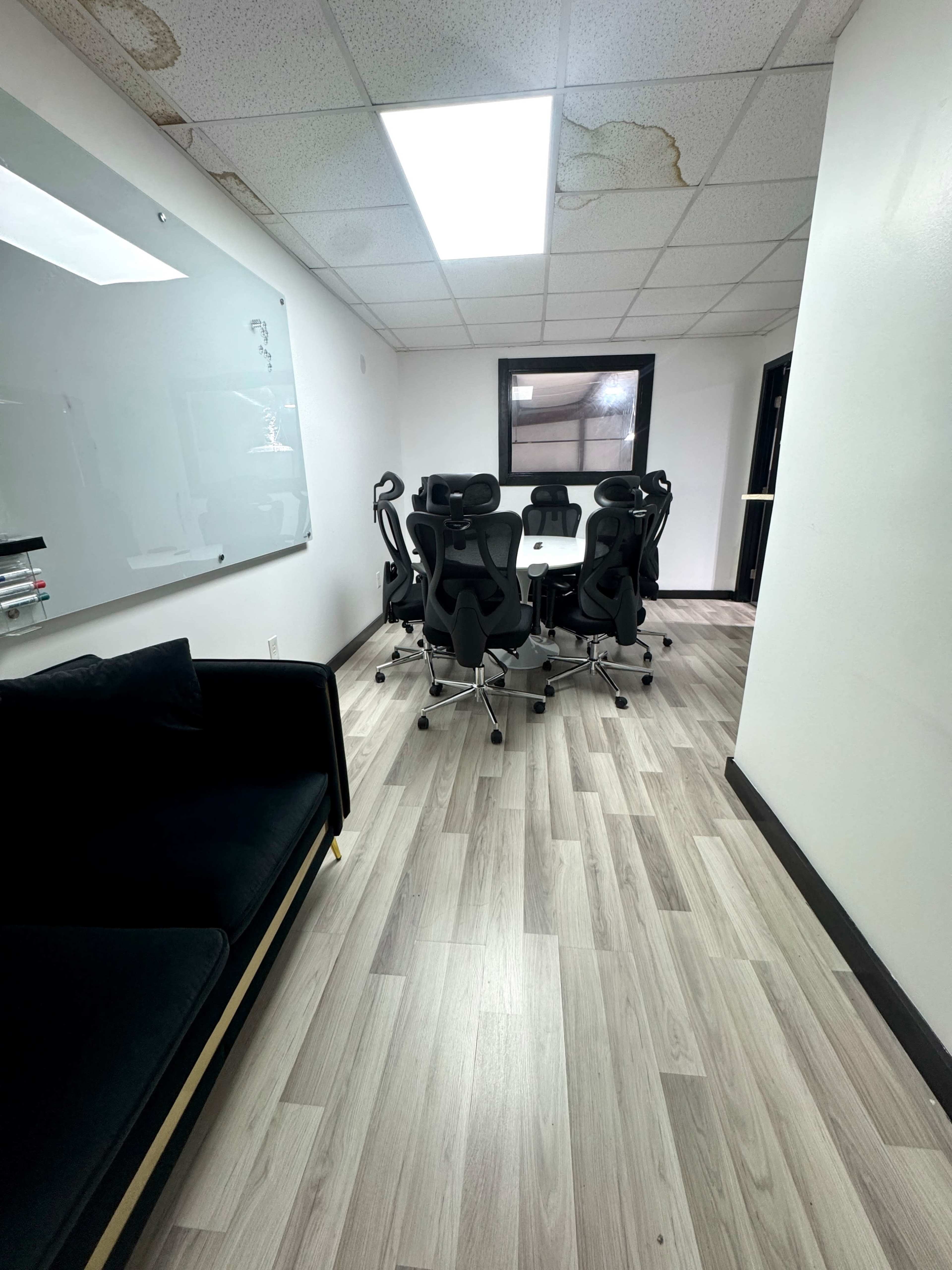 The image shows a conference room with a large whiteboard and a round table surrounded by six black office chairs, along with a dark couch along one wall.