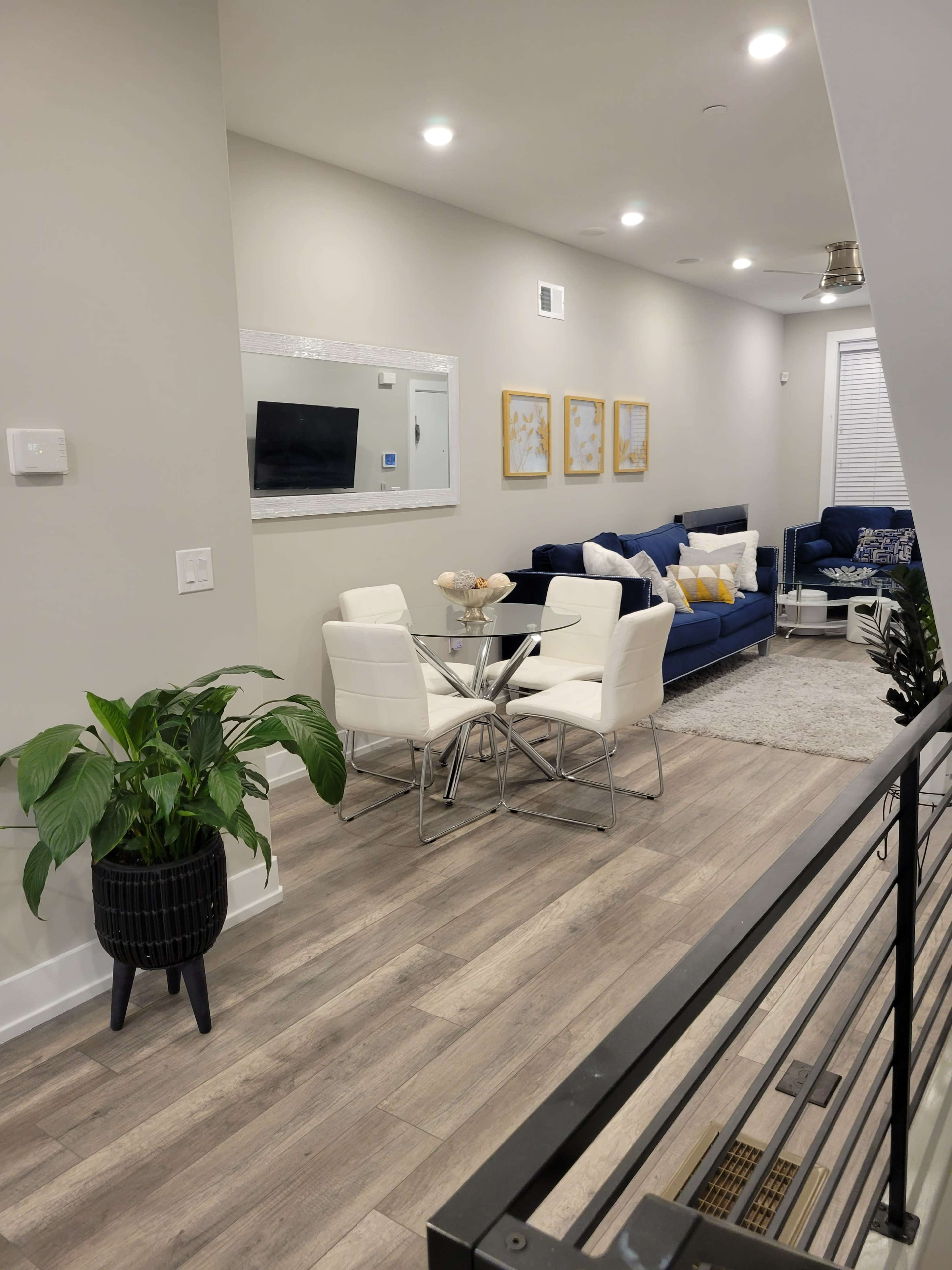 A modern, open living area featuring a glass dining table with four white chairs, a large blue sofa, and decorative framed art on the walls.