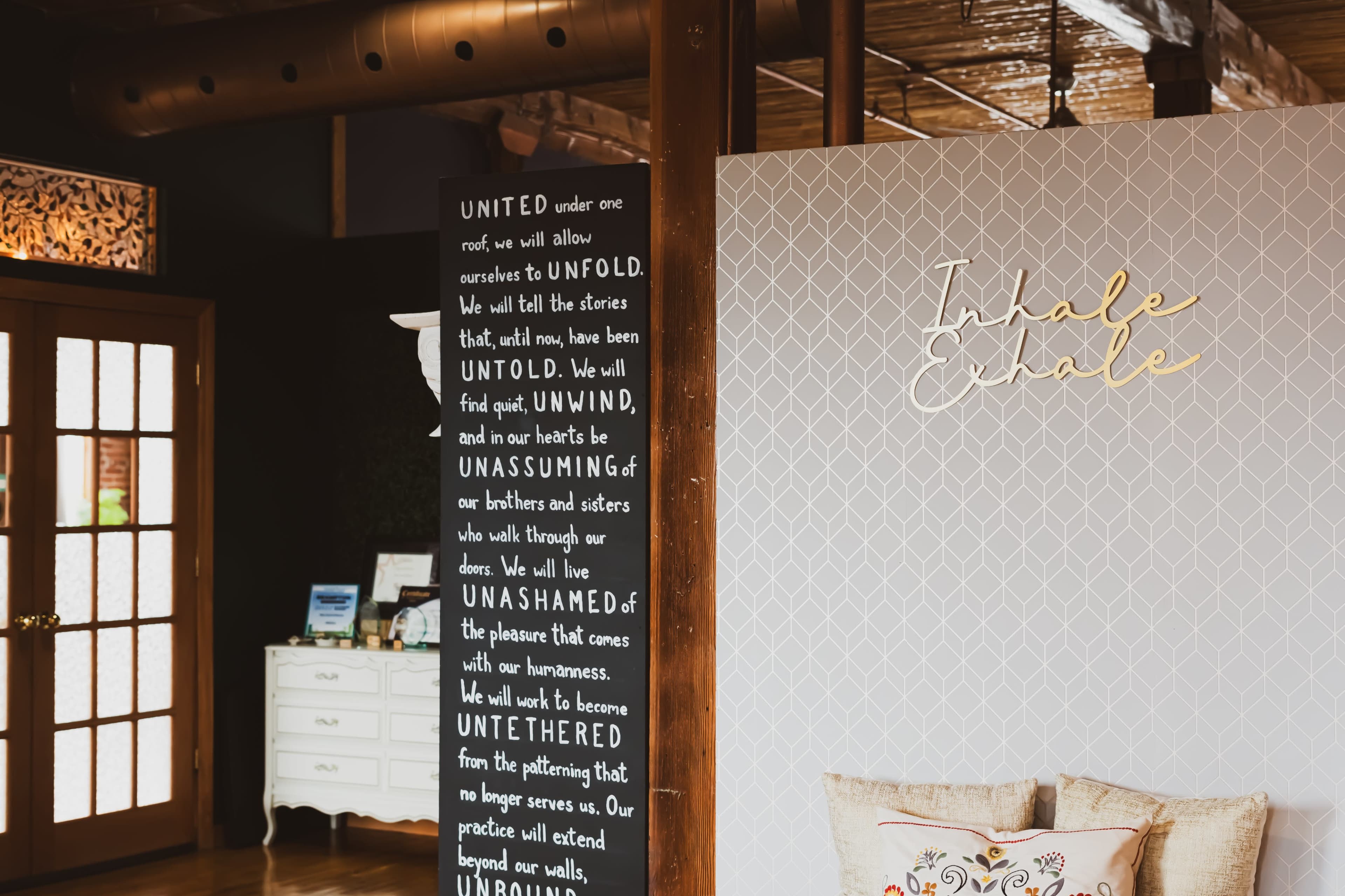 The image shows a cozy interior space featuring a decorative wall with text and another wall with the phrase "Inhale Exhale" illuminated in a warm design.