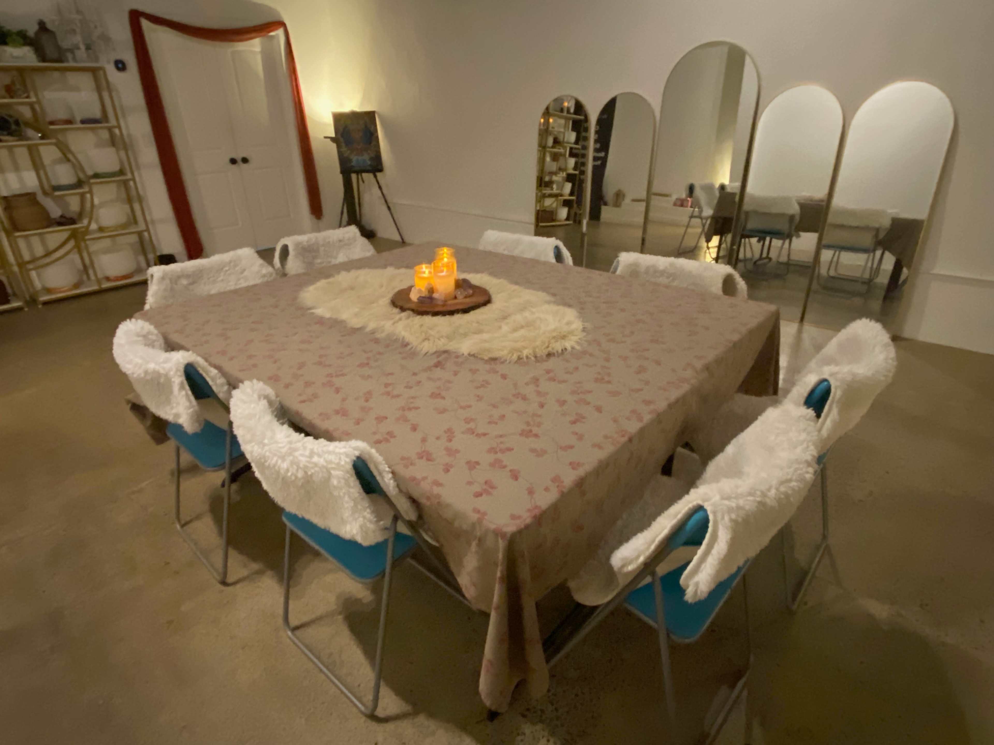 A large rectangular table is set with a floral tablecloth, surrounded by ten chairs, with candles and a decorative centerpiece on top.