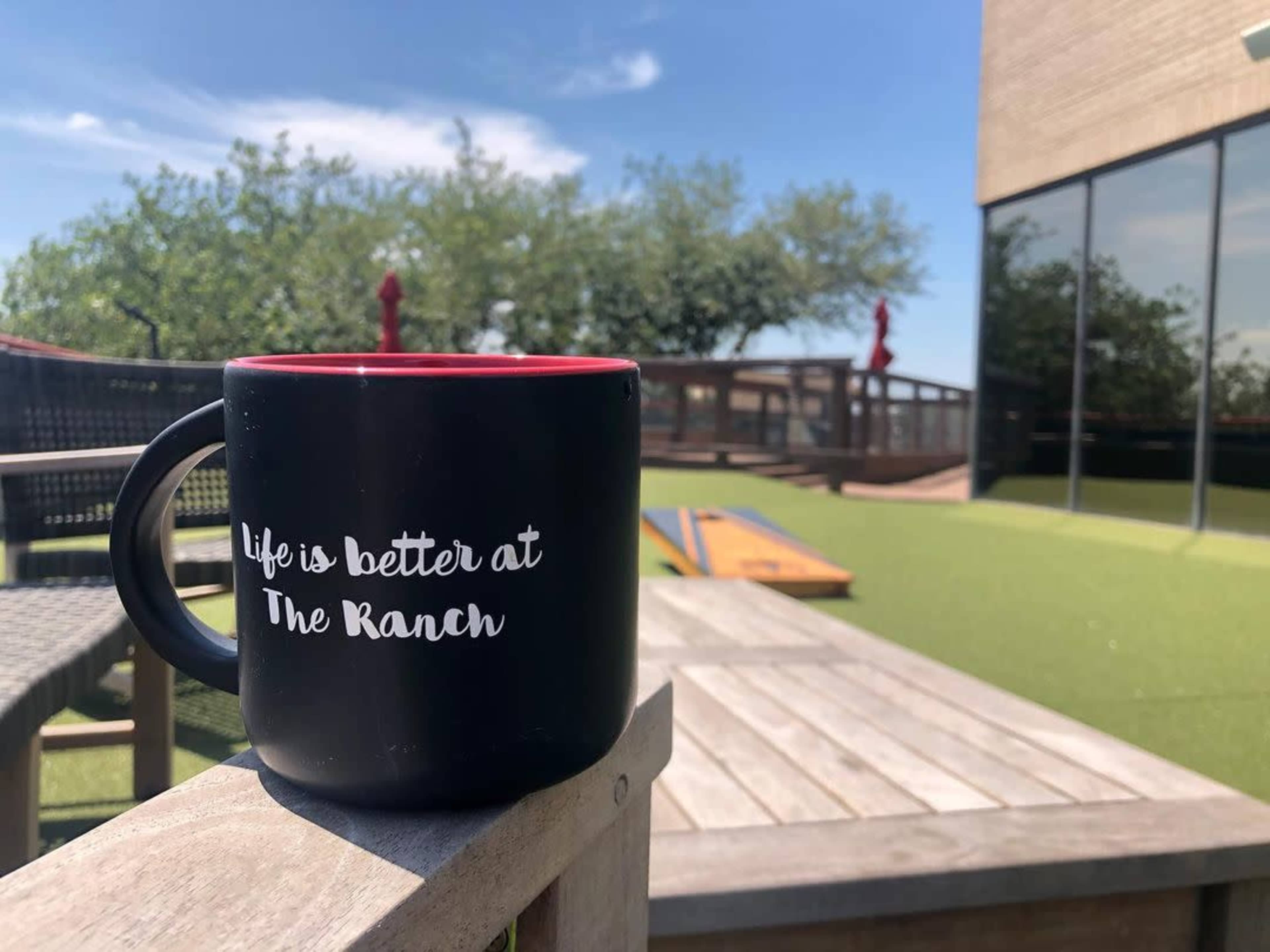 A black coffee mug with a red interior, inscribed with "Life is better at The Ranch," sits on a wooden table in a sunny outdoor area featuring green turf and wooden seating.