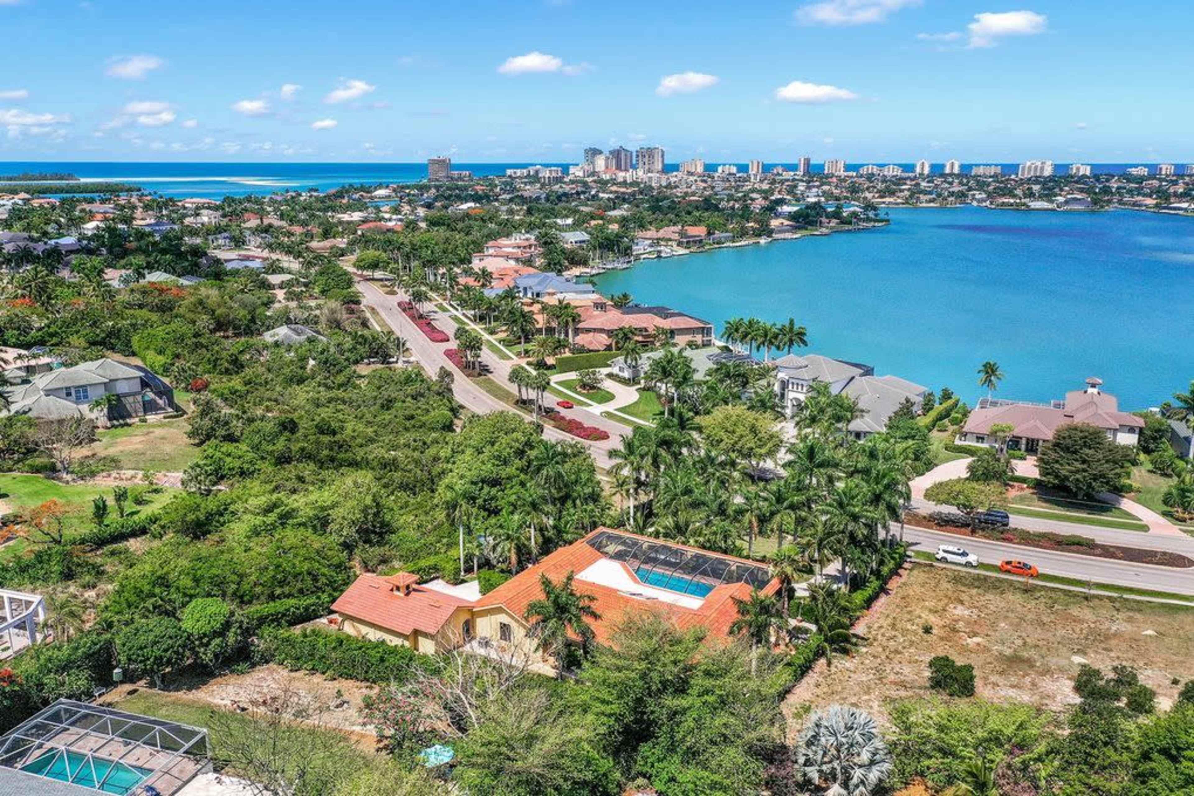 A coastal neighborhood with houses, a swimming pool, and a view of a clear blue bay and distant city skyline.