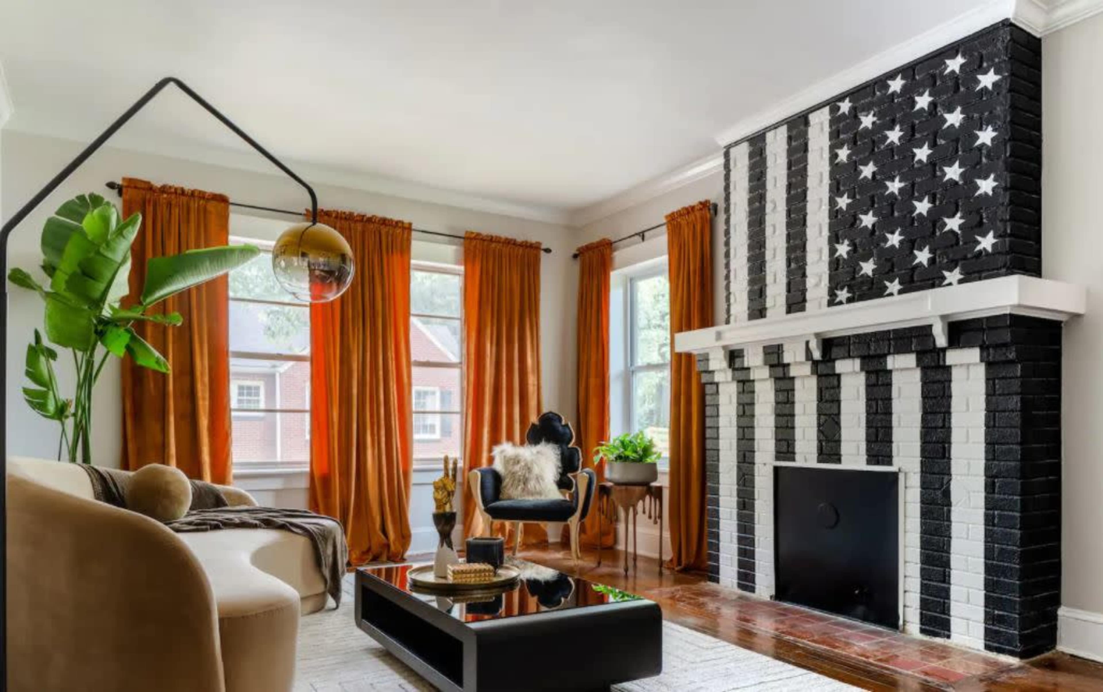 The living room features a black and white striped fireplace adorned with stars, complemented by large orange curtains, a beige sofa, and potted plants.