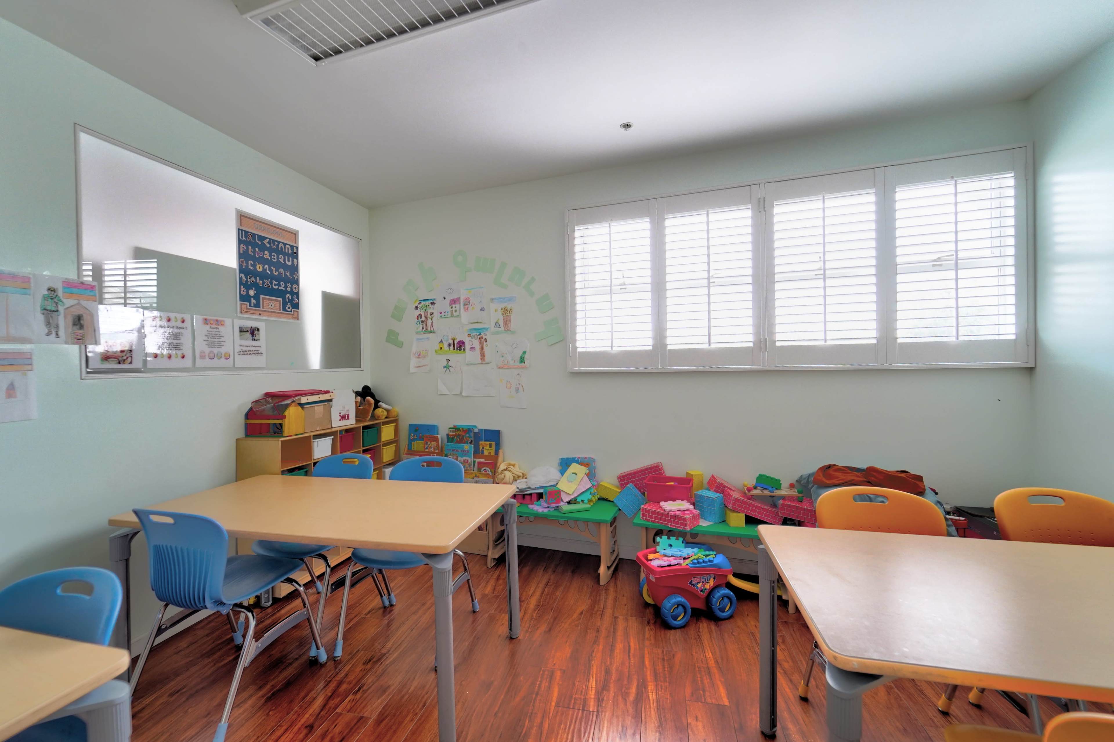 A classroom with several tables and chairs, a toy area with colorful play items, and drawings displayed on the walls.