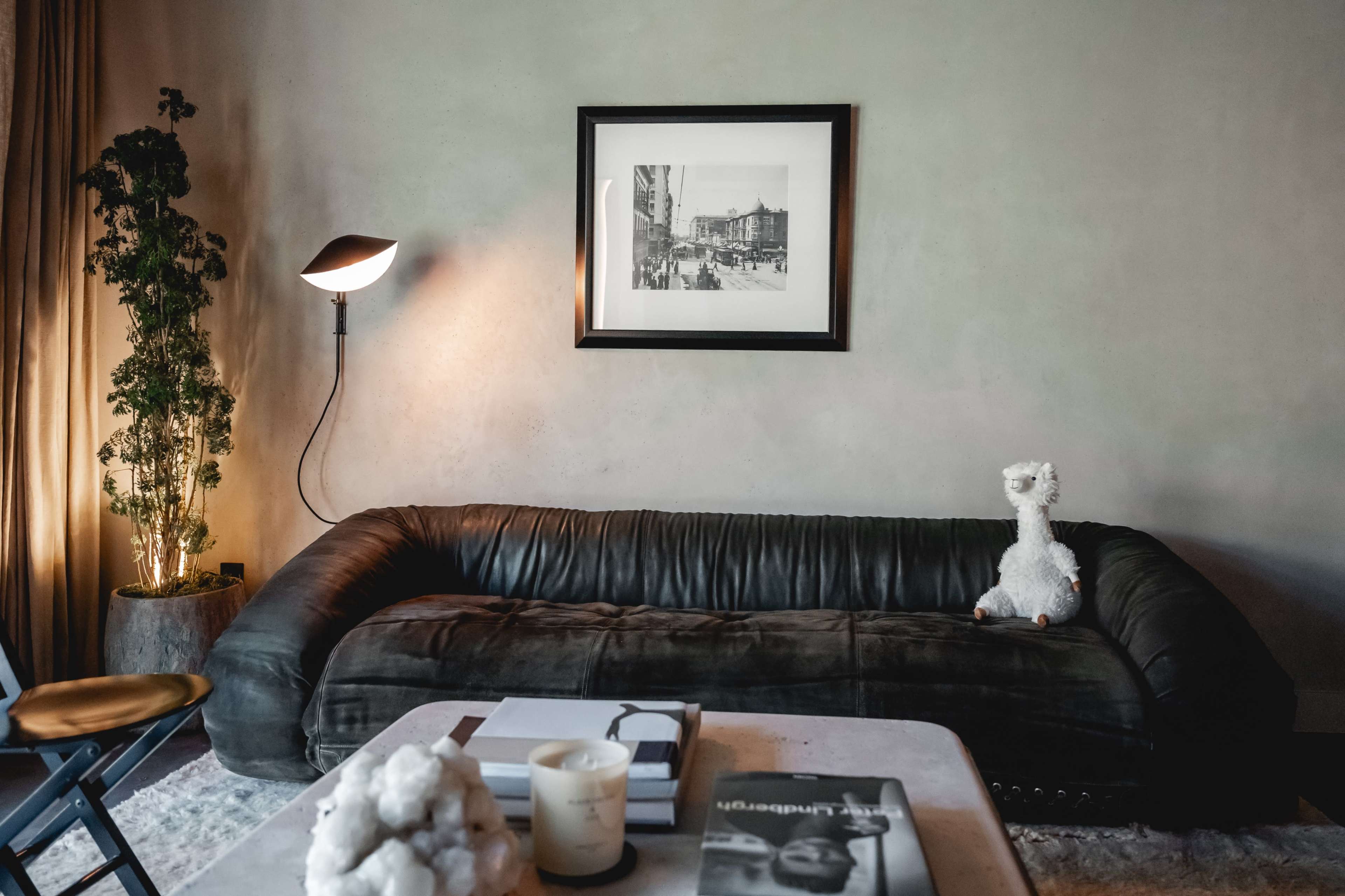 A modern living room with a dark leather couch, a framed black-and-white photograph on the wall, a light fixture, and a decorative white stuffed animal on the couch.