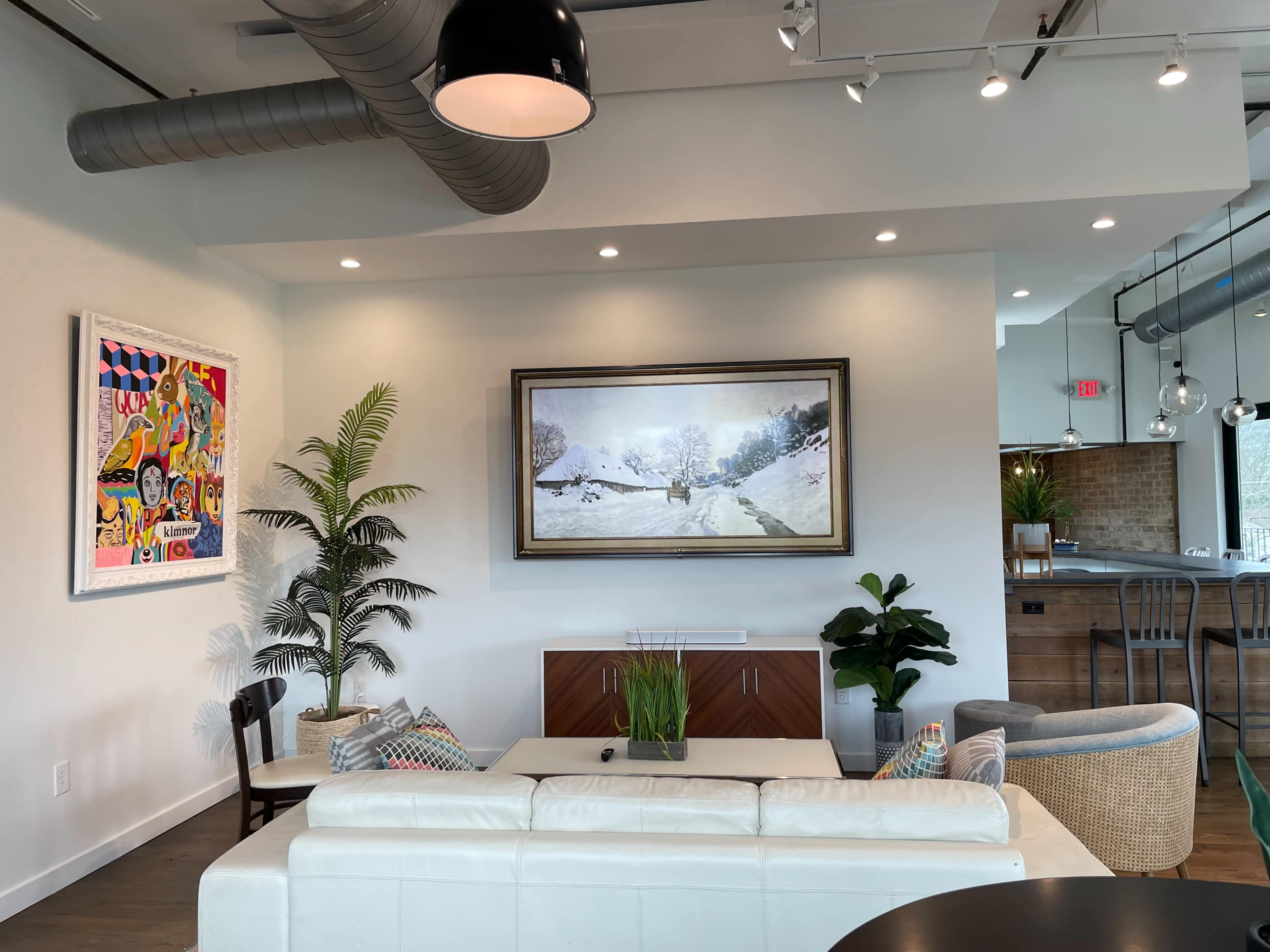 A modern interior space featuring a white sofa, a large framed painting on the wall, colorful artwork, and various plants.