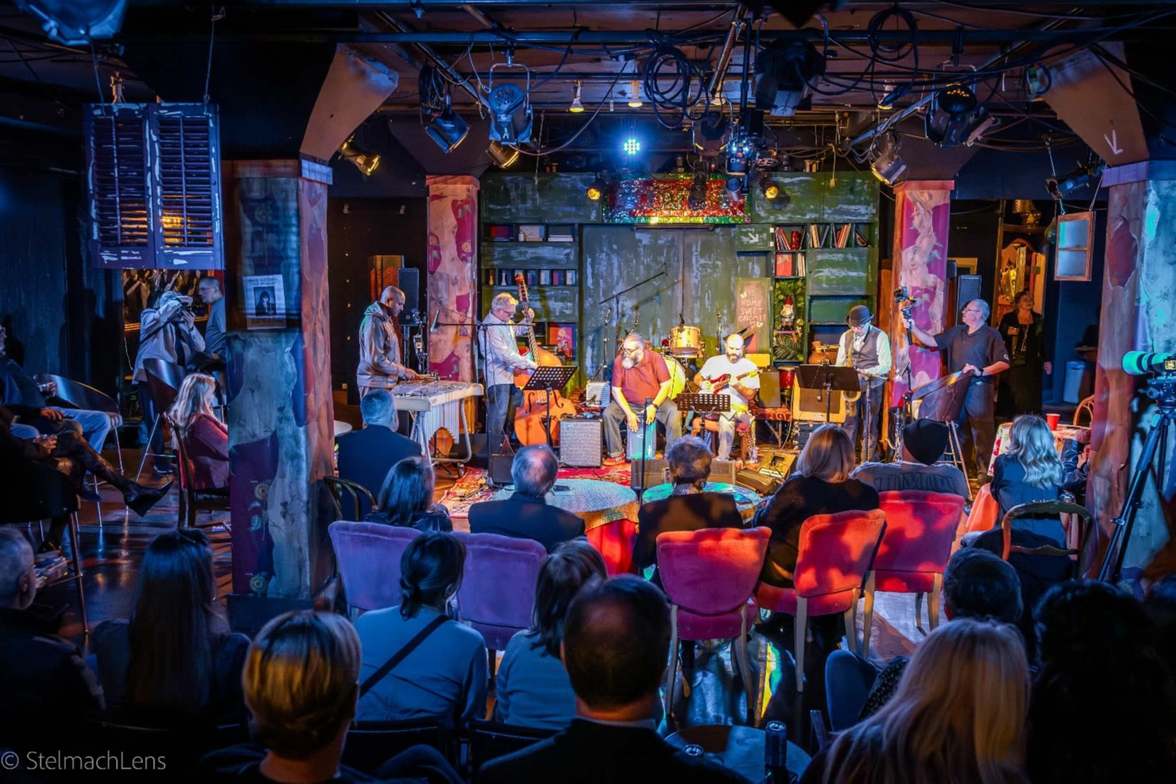 A group of musicians performs on stage in a dimly lit venue, while an audience watches from chairs arranged in front.