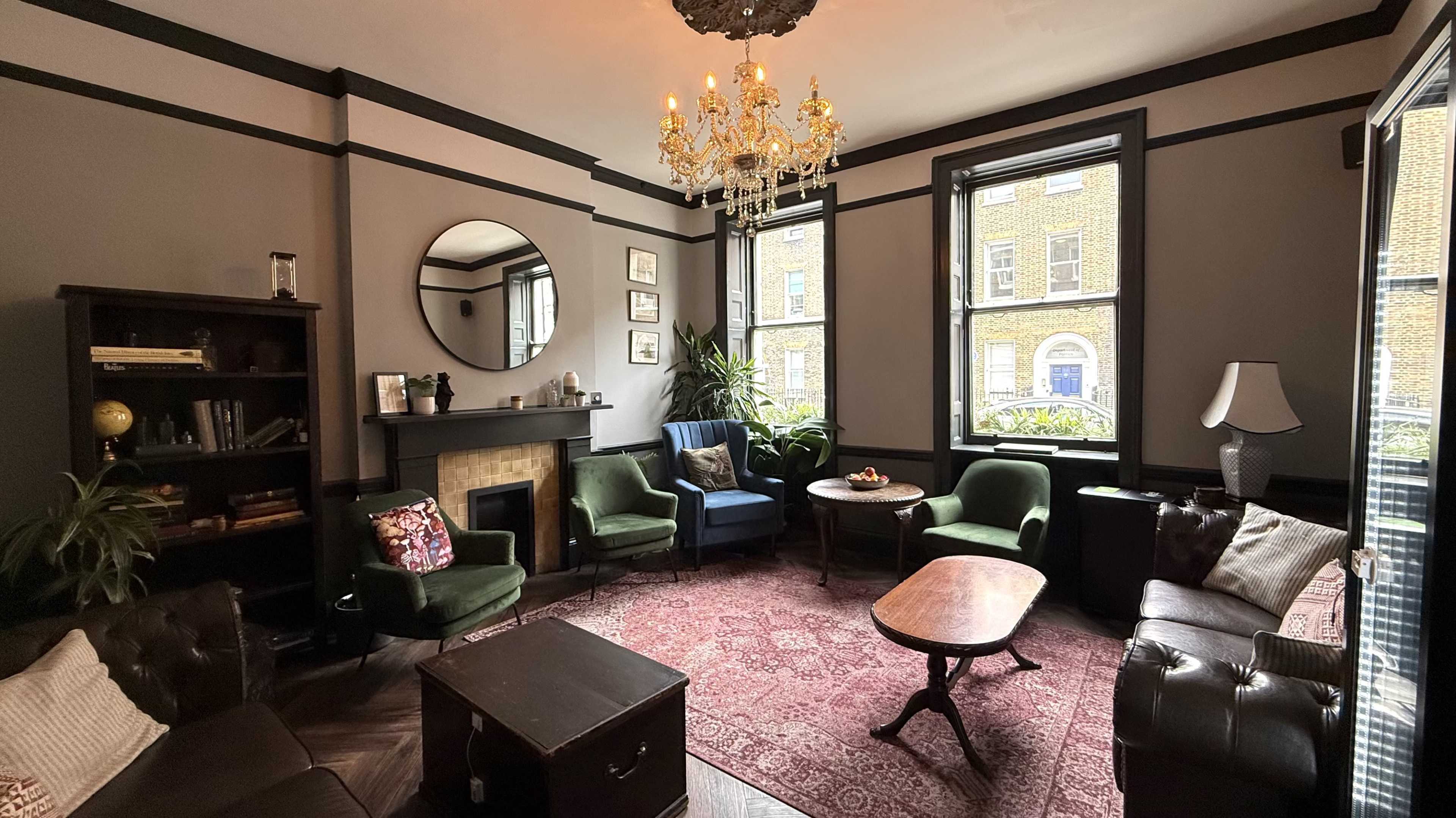 The image shows a cozy living room with a mix of seating options, including green armchairs and a brown leather couch, surrounded by windows and decorated with a chandelier and bookshelf.