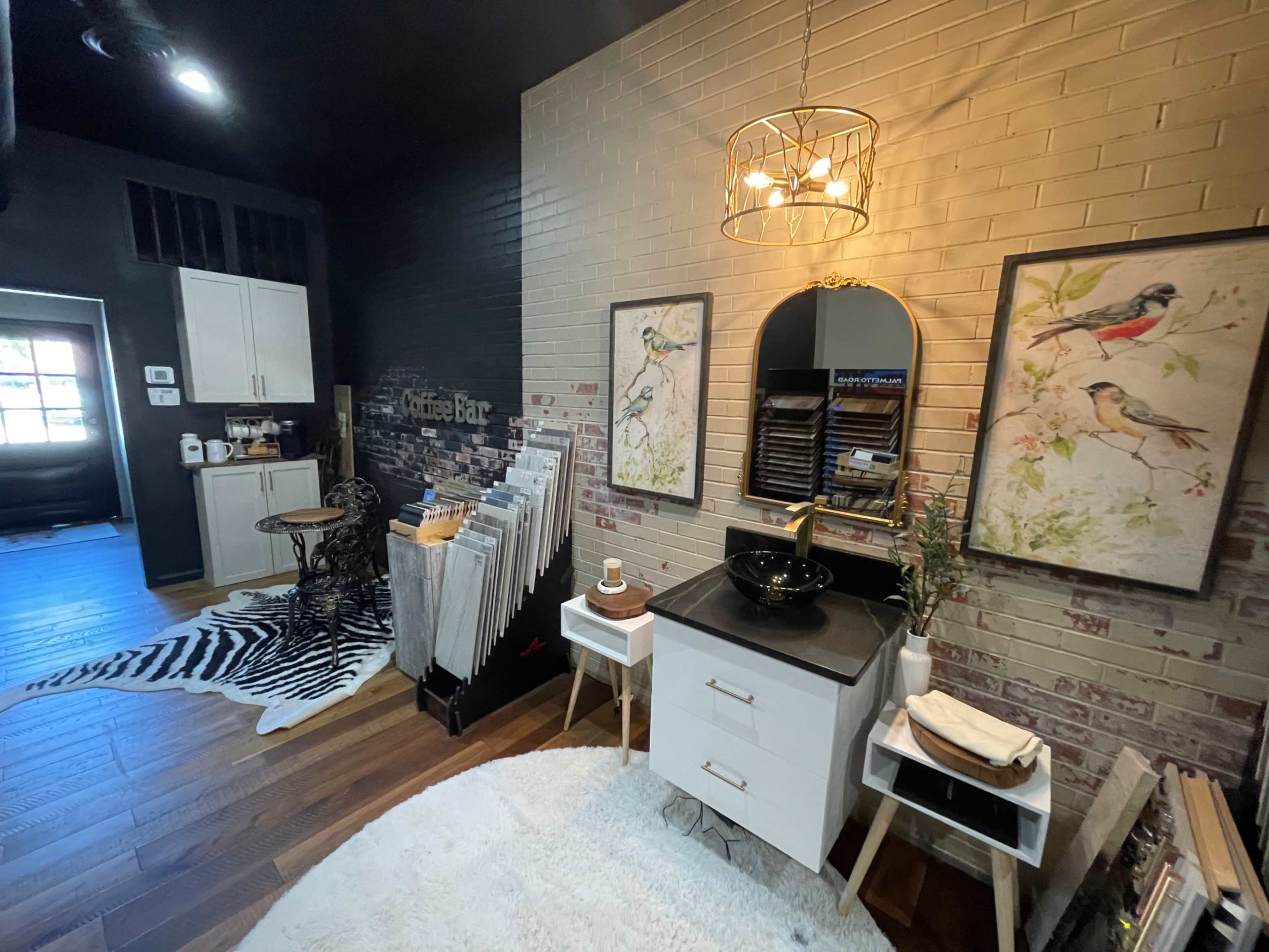 A stylish interior space featuring a wall with brick accents, a mirror above a small black sink, framed bird paintings, and a collection of magazines displayed on a wooden stand.