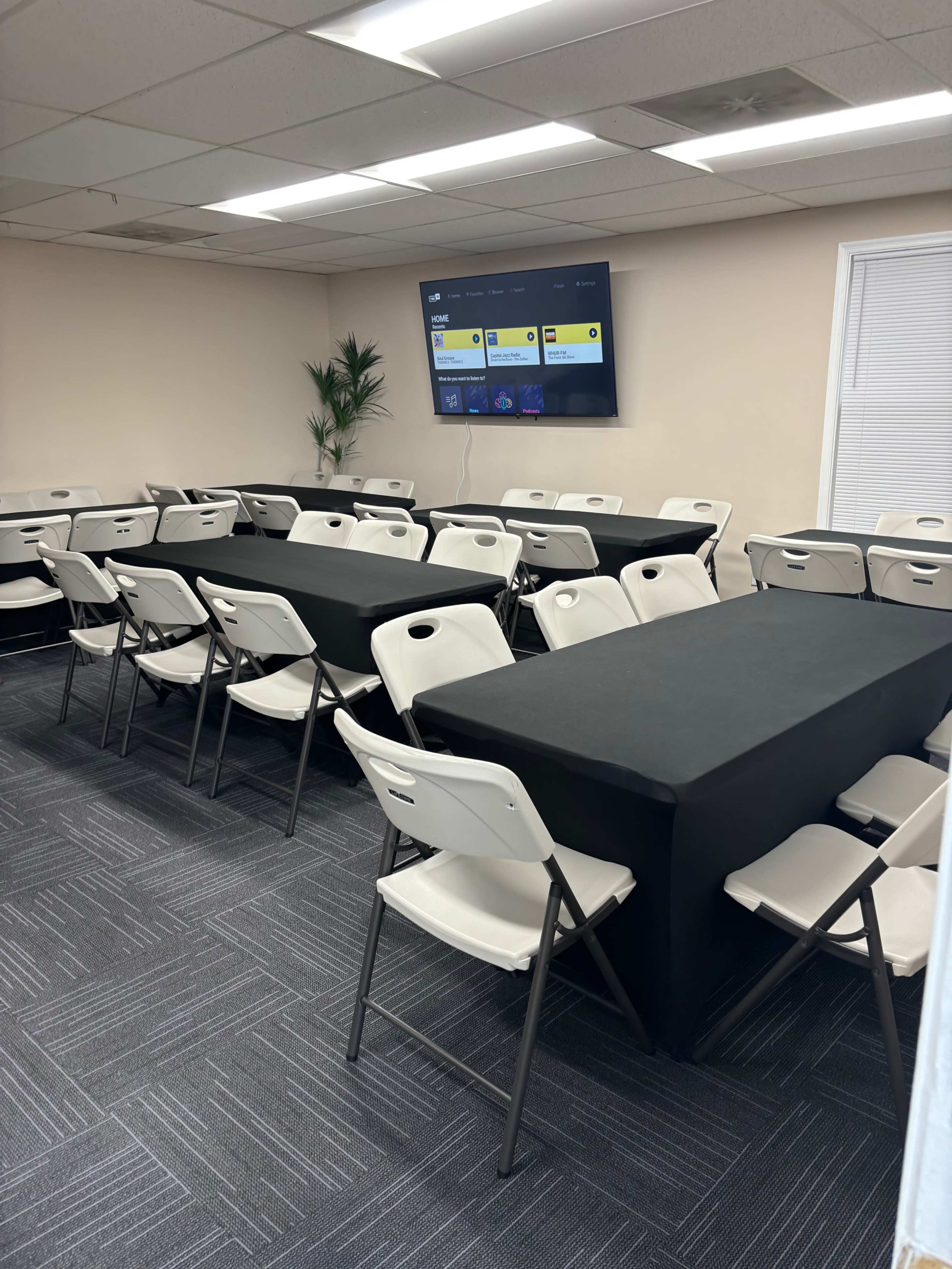 A conference room features several rows of chairs arranged around black-covered tables, with a television mounted on the wall displaying a screen interface.