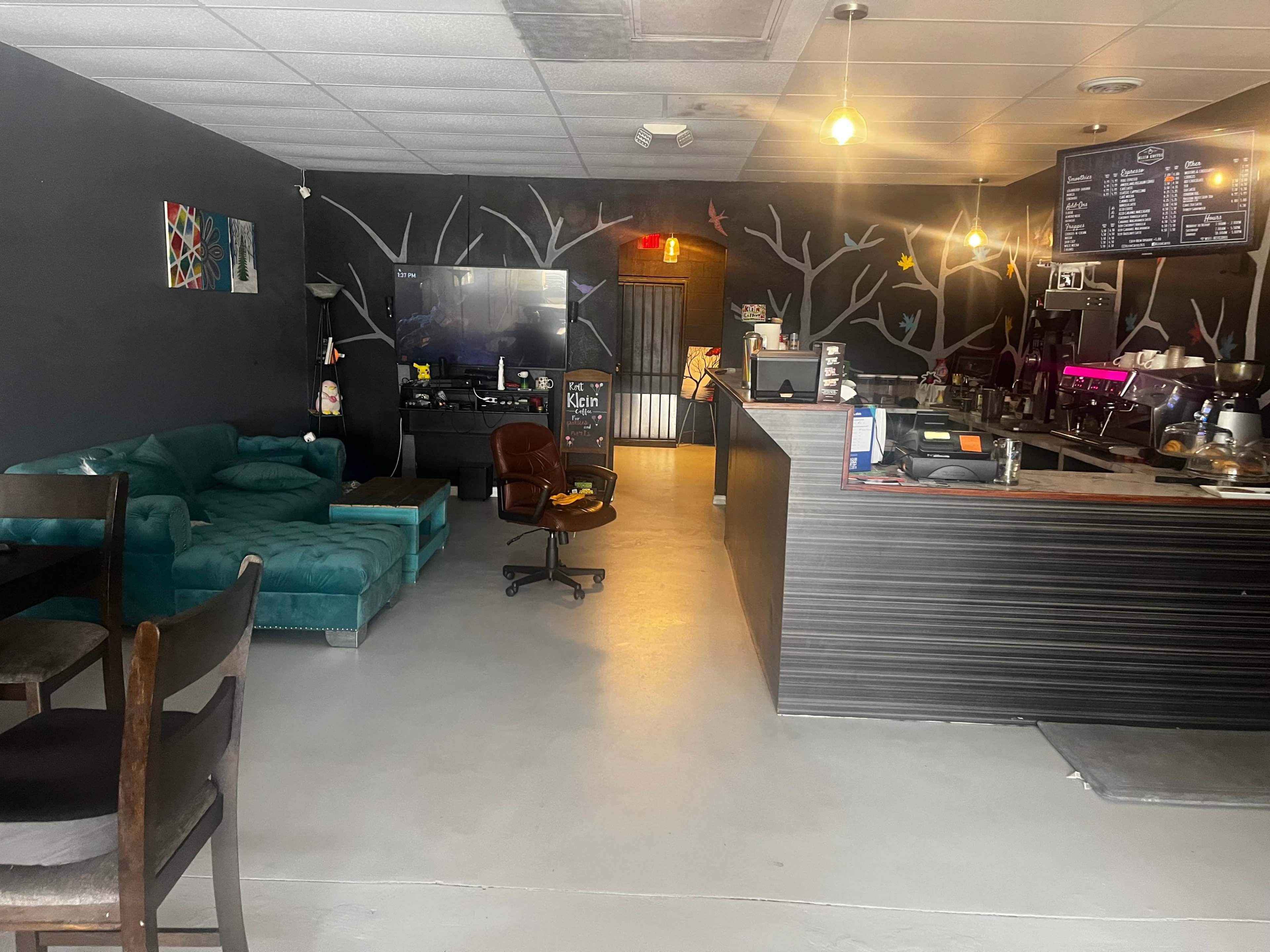A cozy café interior with a teal couch, wooden tables, and a counter displaying a menu, set against wall murals of trees.
