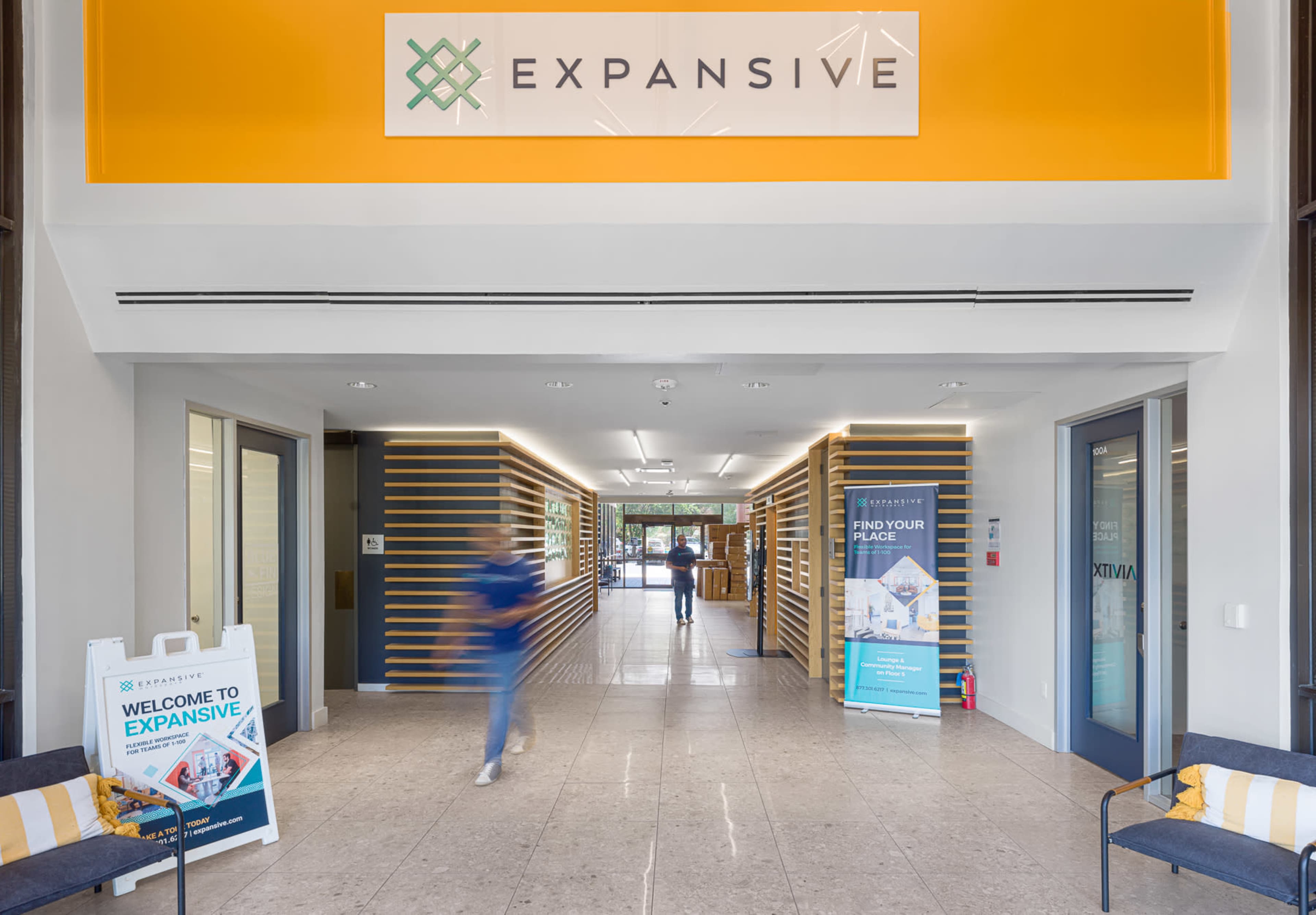 A modern office entrance features a sign that reads "EXPANSIVE" above a spacious corridor with seating on either side.