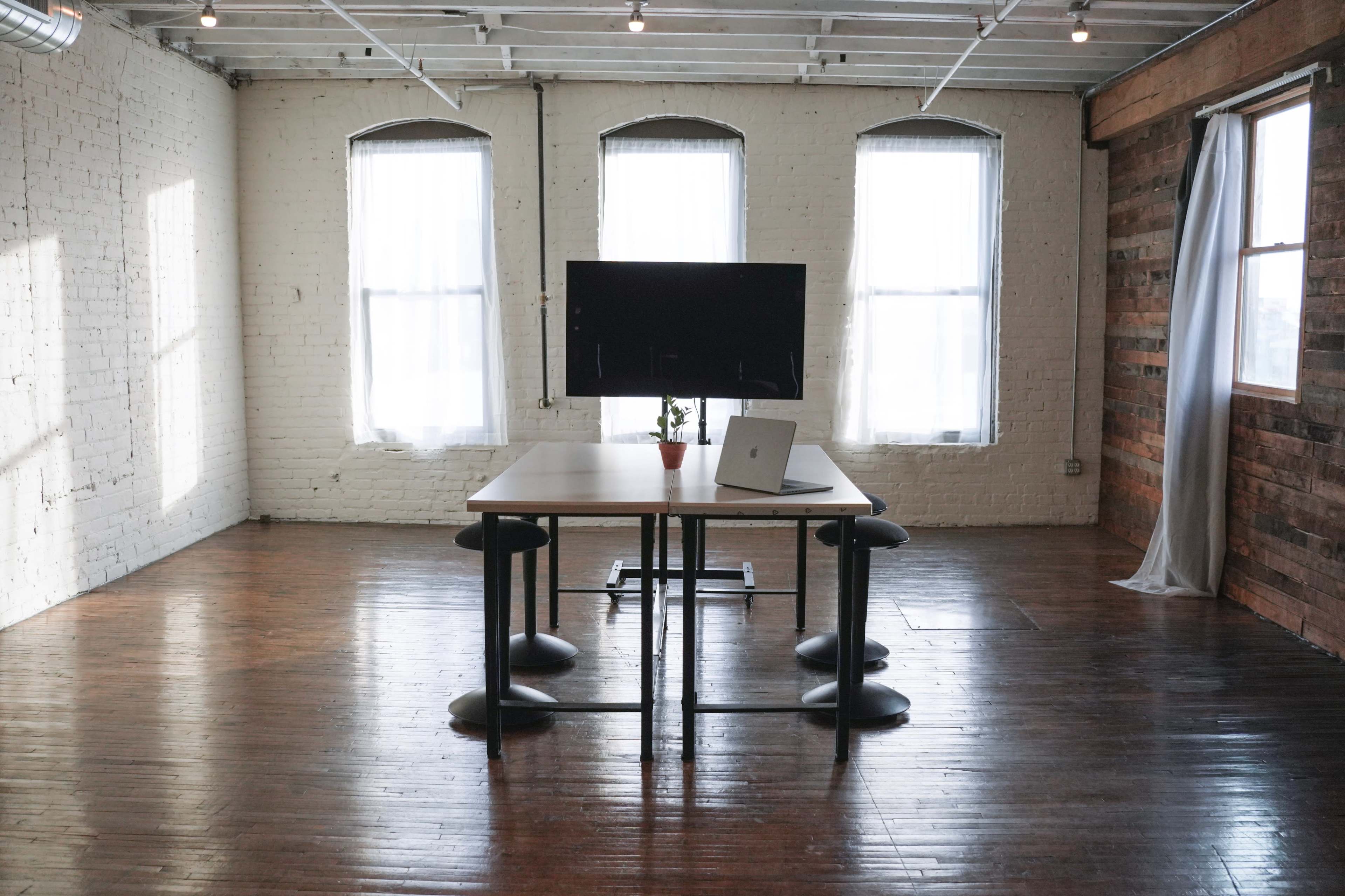 A modern workspace with a large table, four black stools, a computer on the table, and large windows allowing natural light to fill the room.