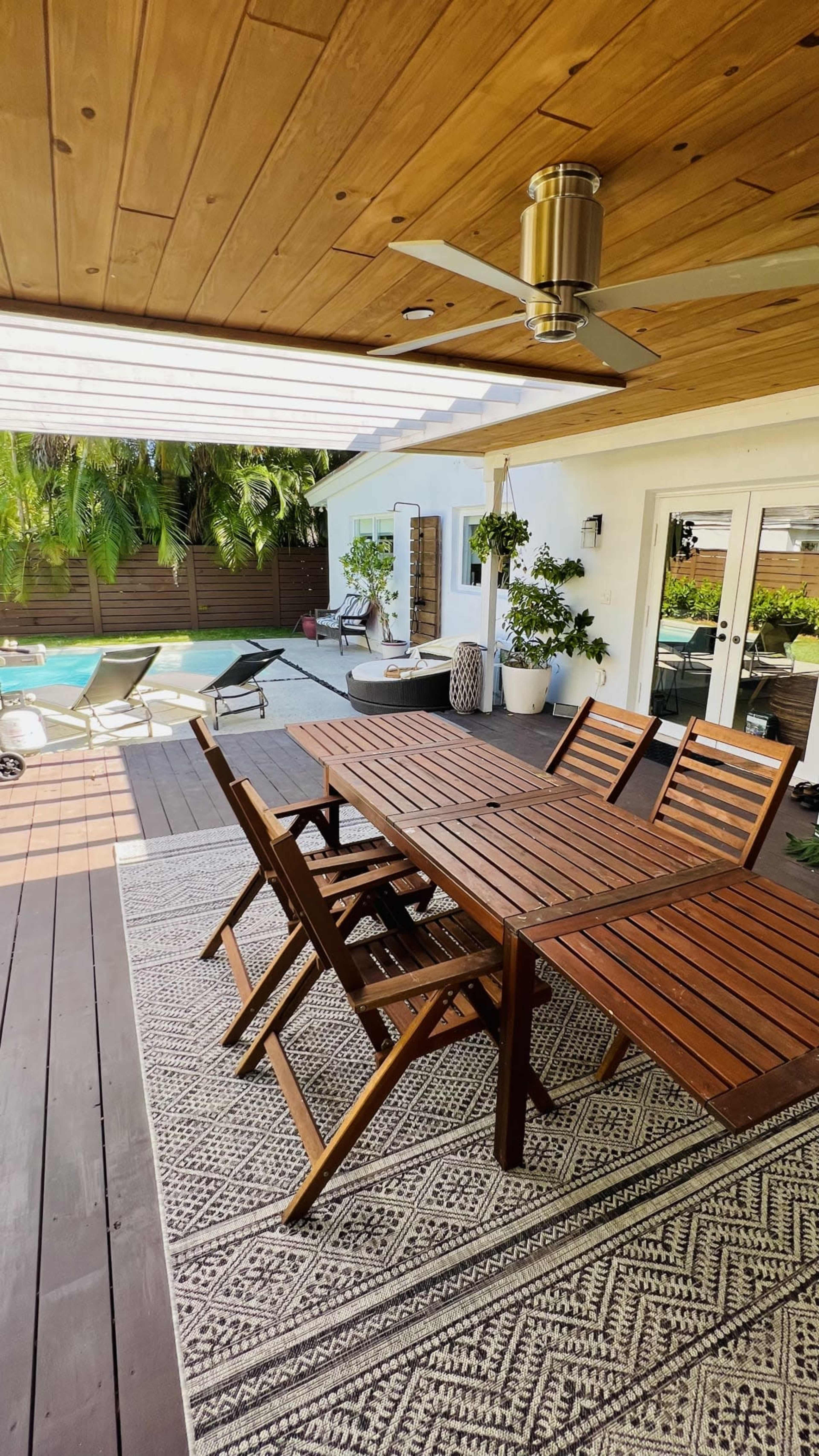 A wooden dining table with matching chairs is positioned on a deck that overlooks a swimming pool surrounded by tropical plants.
