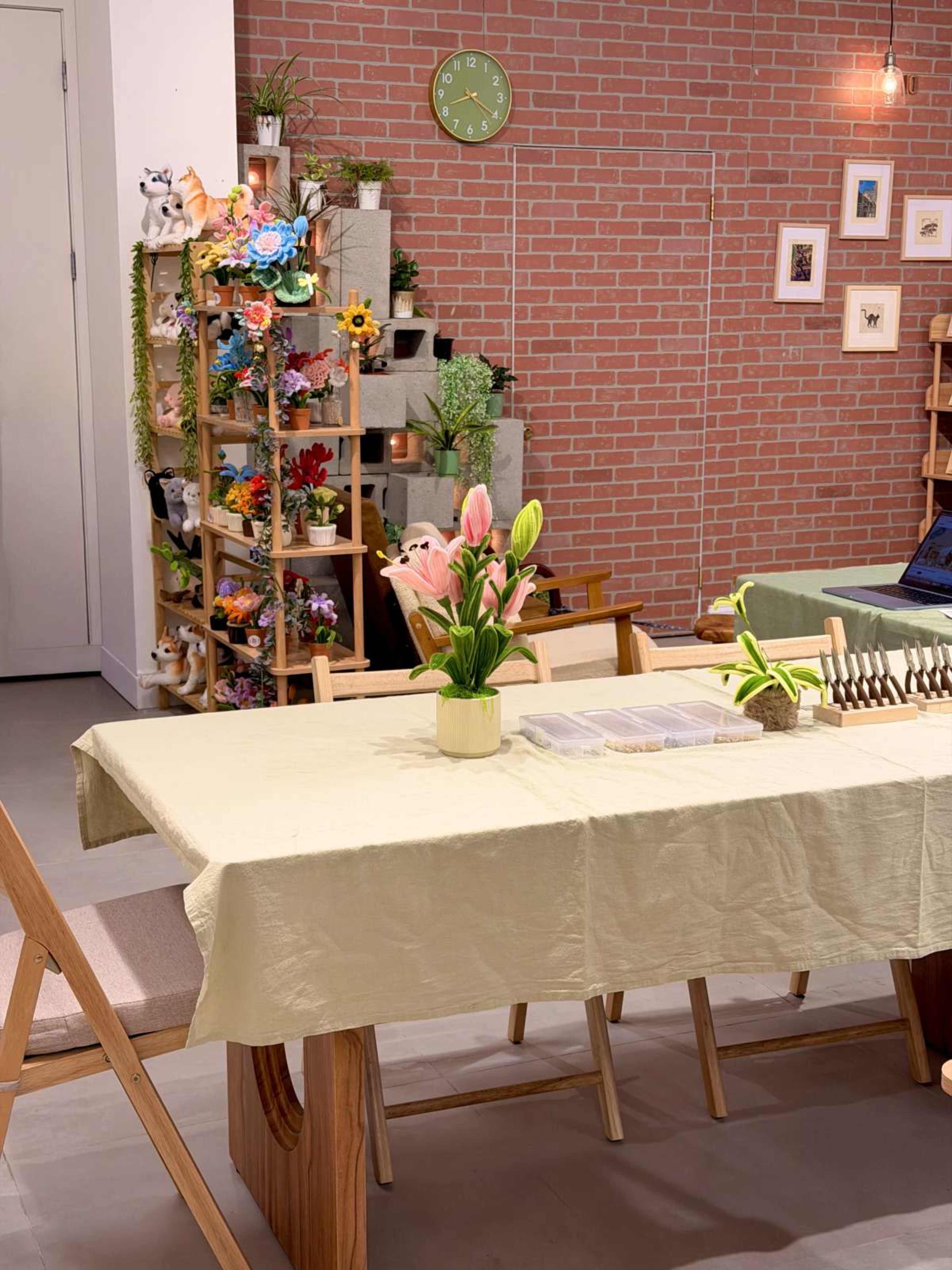 The image shows a cozy indoor space with a wooden table set for a meal, a vase of lilies in the center, and a decorative shelf filled with plants and ornamental items against a brick wall.