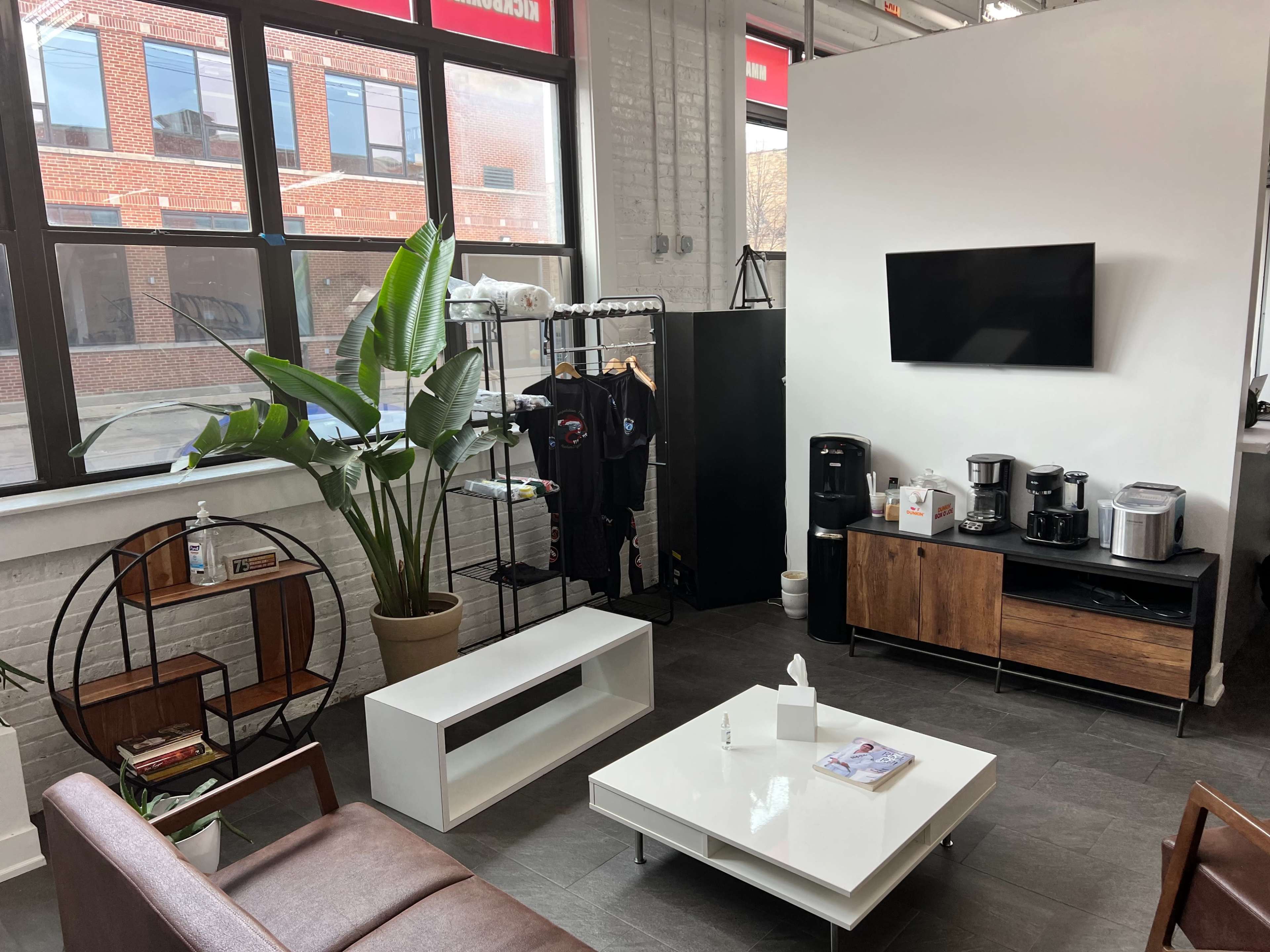 A modern office lounge area with a coffee station, a white coffee table, a brown sofa, and large windows letting in natural light.