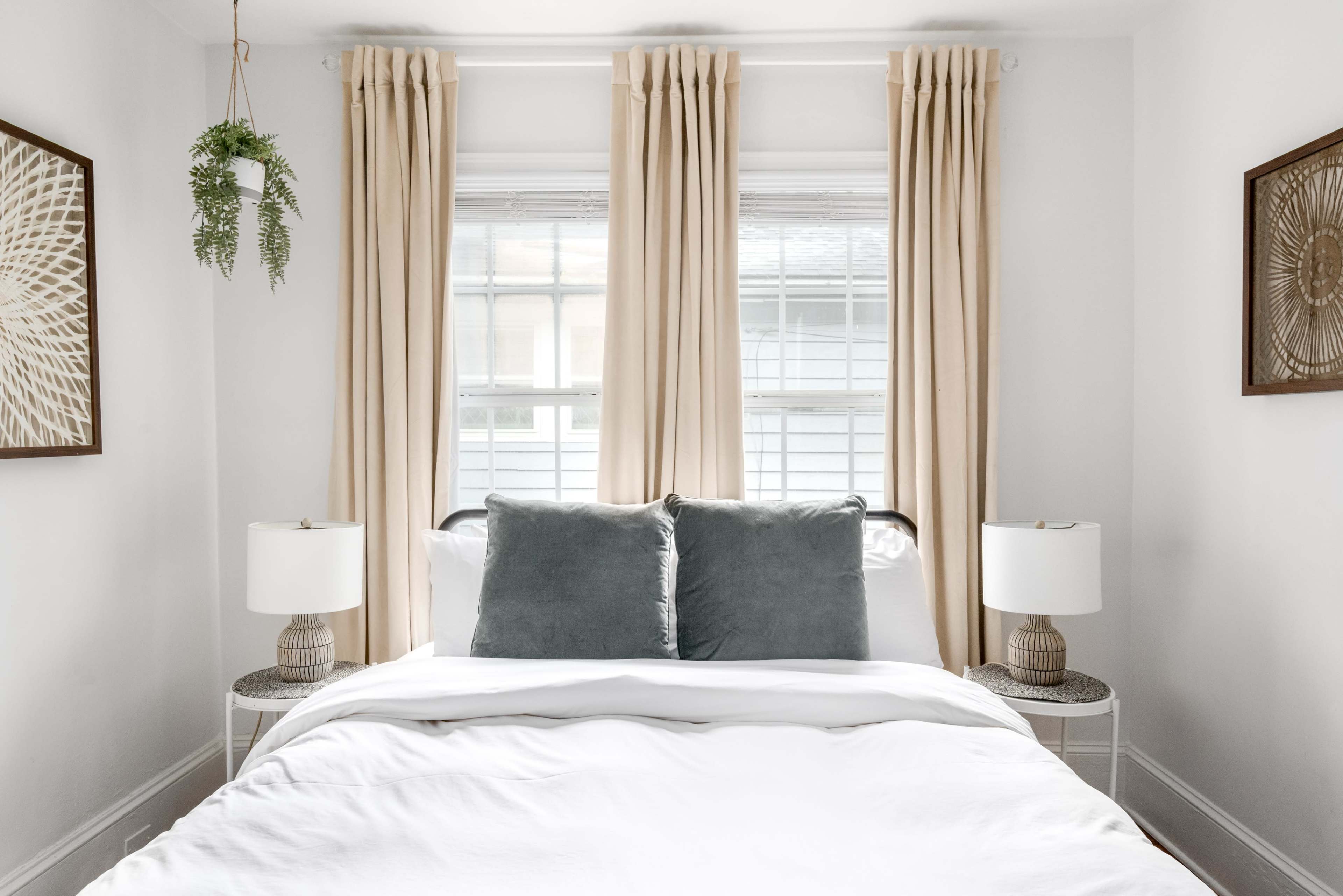 A neatly made bed with gray cushions is framed by beige curtains and accompanied by two bedside lamps in a bright, minimalist bedroom.