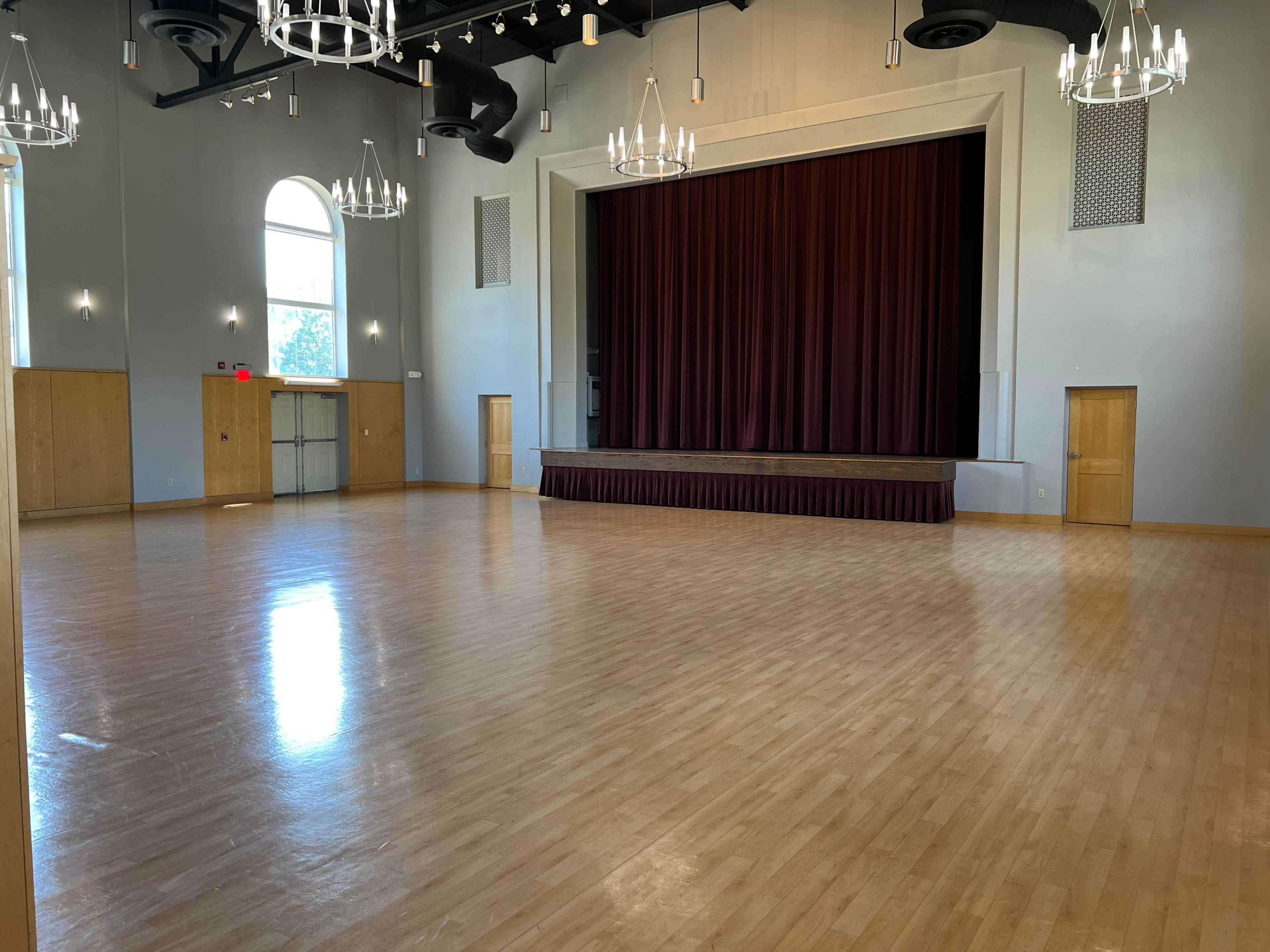 A spacious indoor hall with a polished wooden floor, a stage at the front, and large windows allowing natural light to enter.
