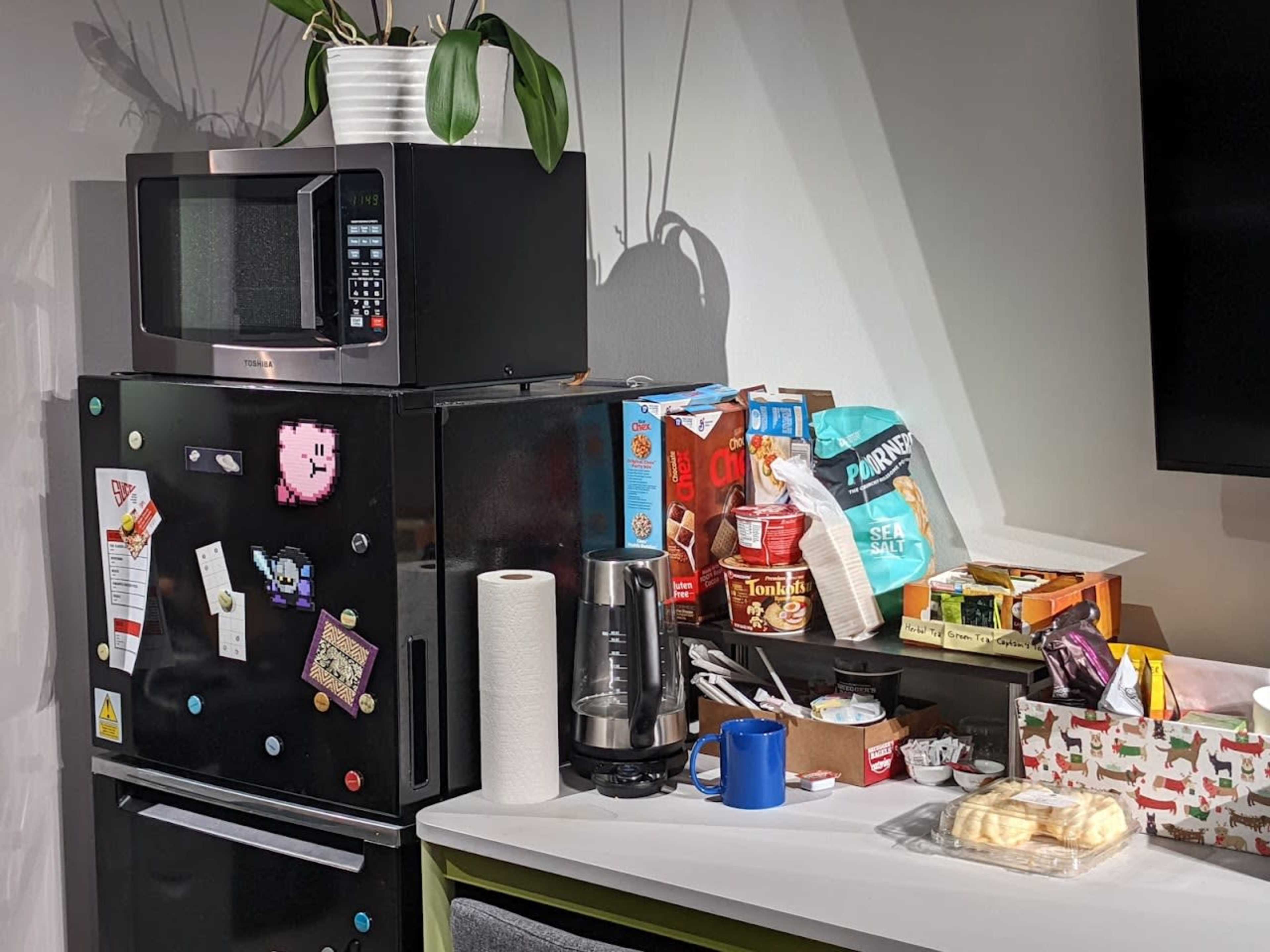 A kitchen counter with a black refrigerator, a microwave on top, and various food items and utensils scattered on the counter.