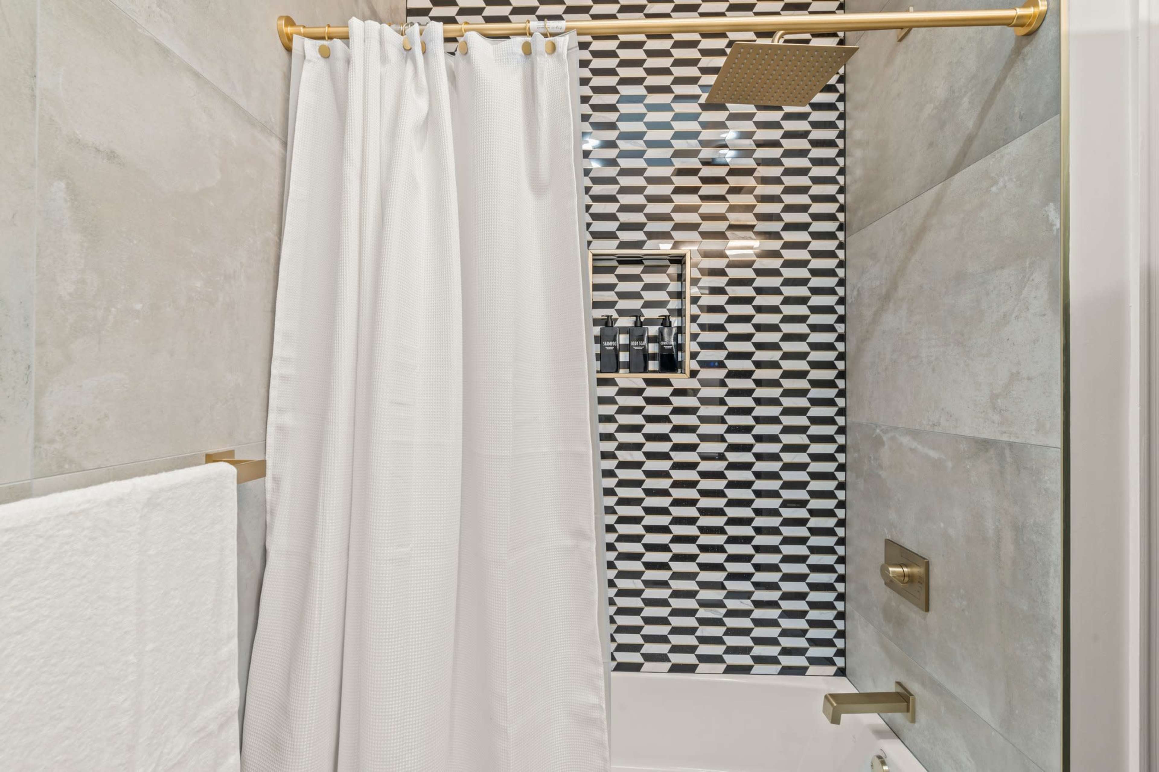 The image shows a modern bathroom with a white shower curtain, a tiled wall featuring a black-and-white geometric pattern, and a gold shower fixture.