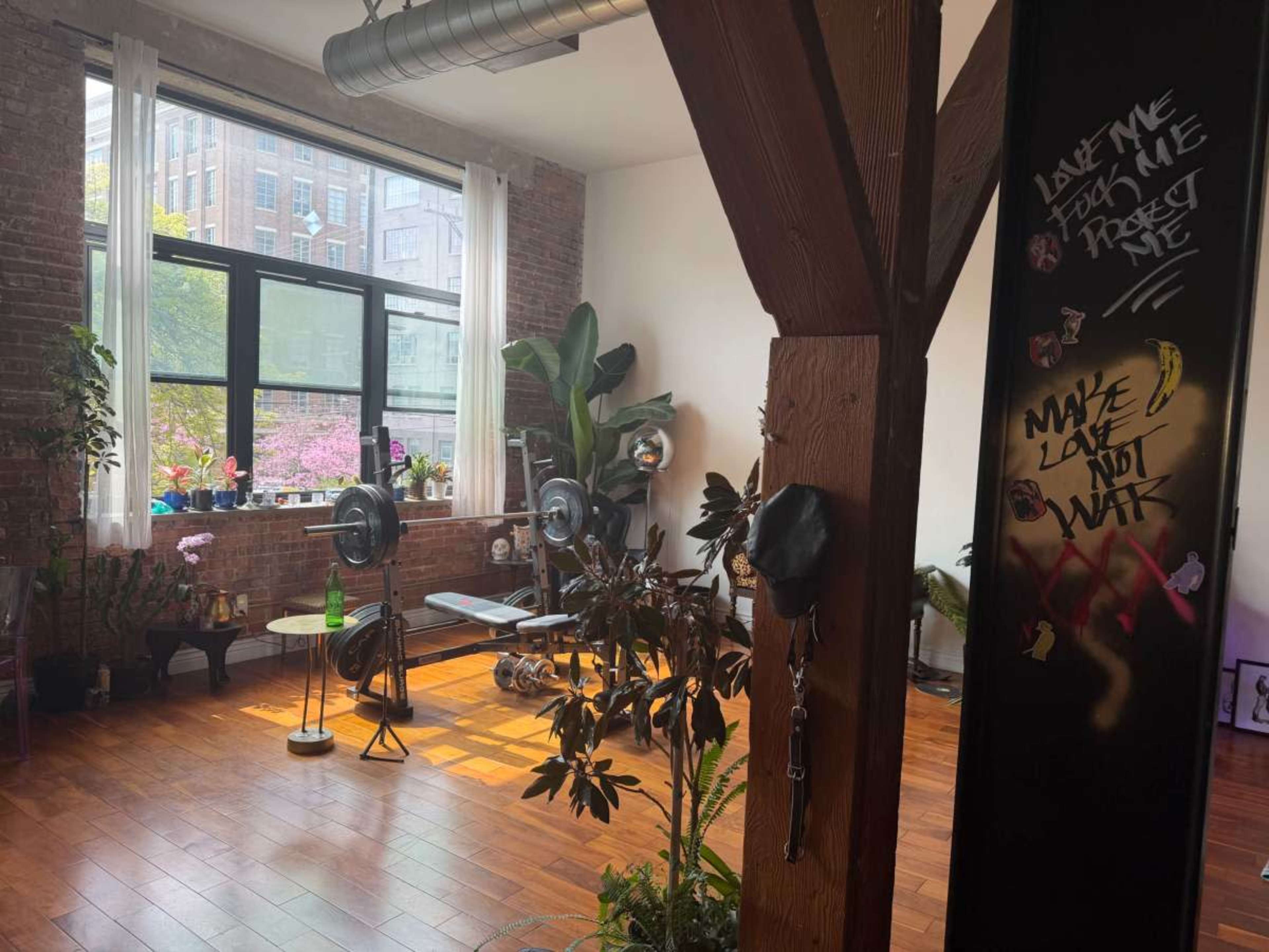 The image shows an indoor gym space with a weight bench, weights, and plants, located in a room with large windows and exposed brick walls.
