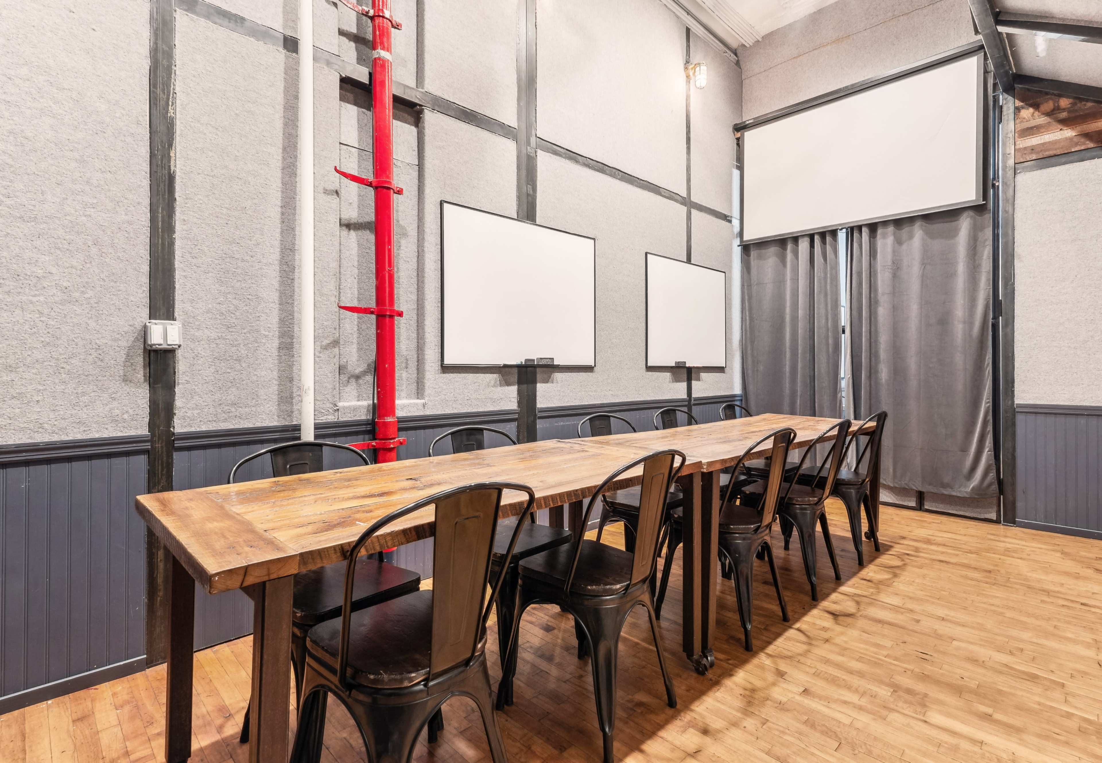 A meeting room features a long wooden table surrounded by black chairs, with gray walls and two white screens at the front.